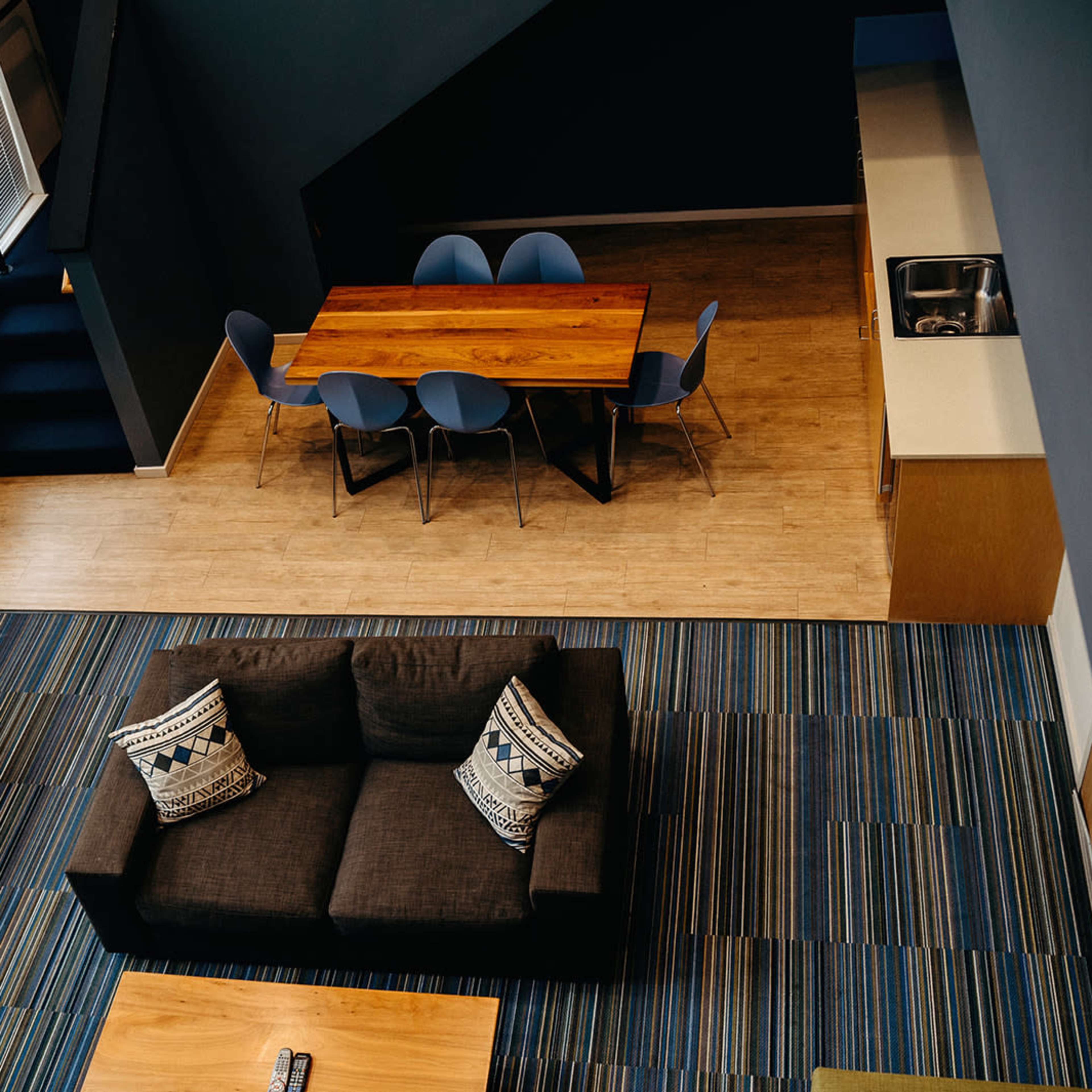A modern living space featuring a dark sofa with decorative pillows, a wooden dining table surrounded by blue chairs, and a kitchen area, all set against a backdrop of striped flooring.