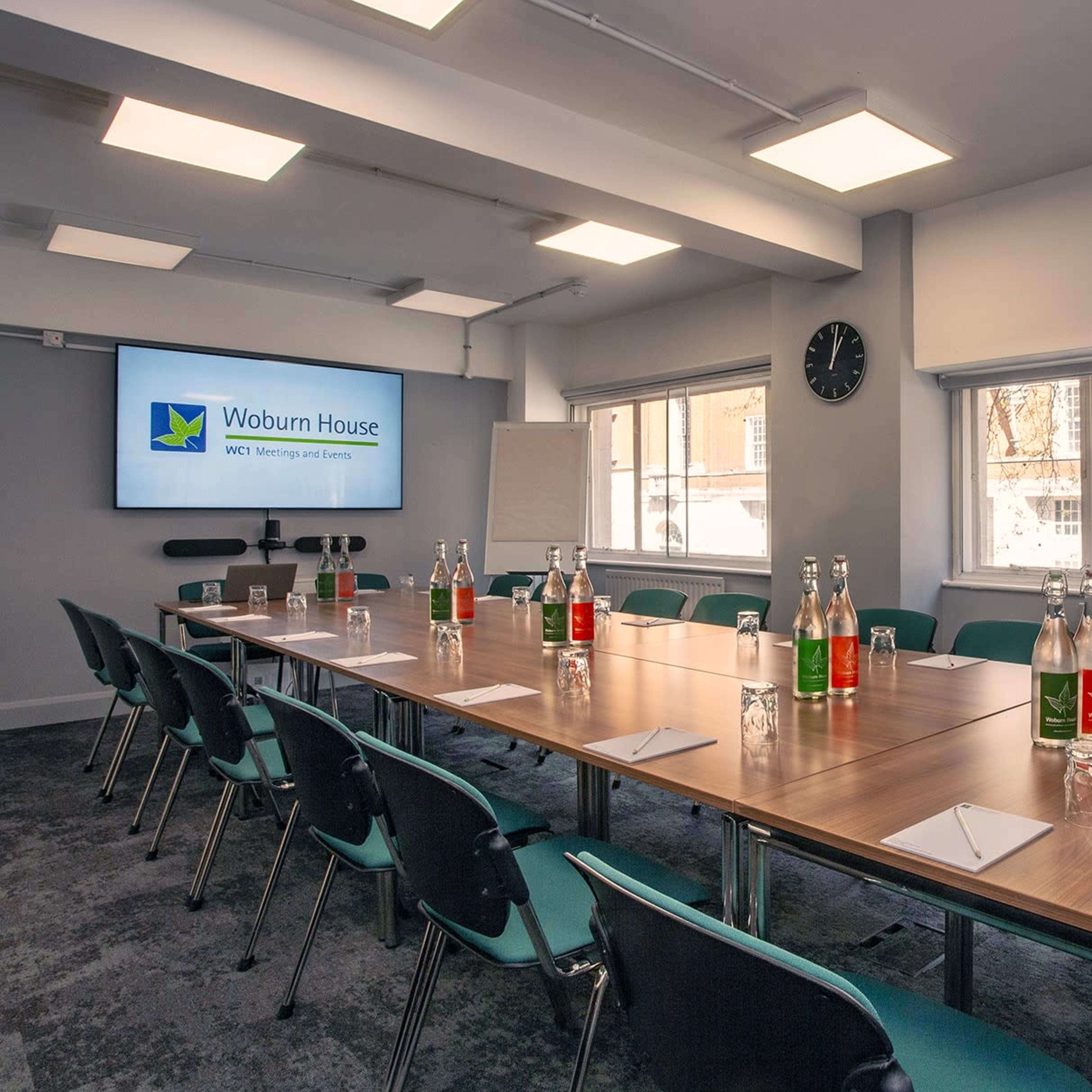 A meeting room with a large rectangular table set for a conference, featuring water bottles, notepads, and a presentation screen at one end.