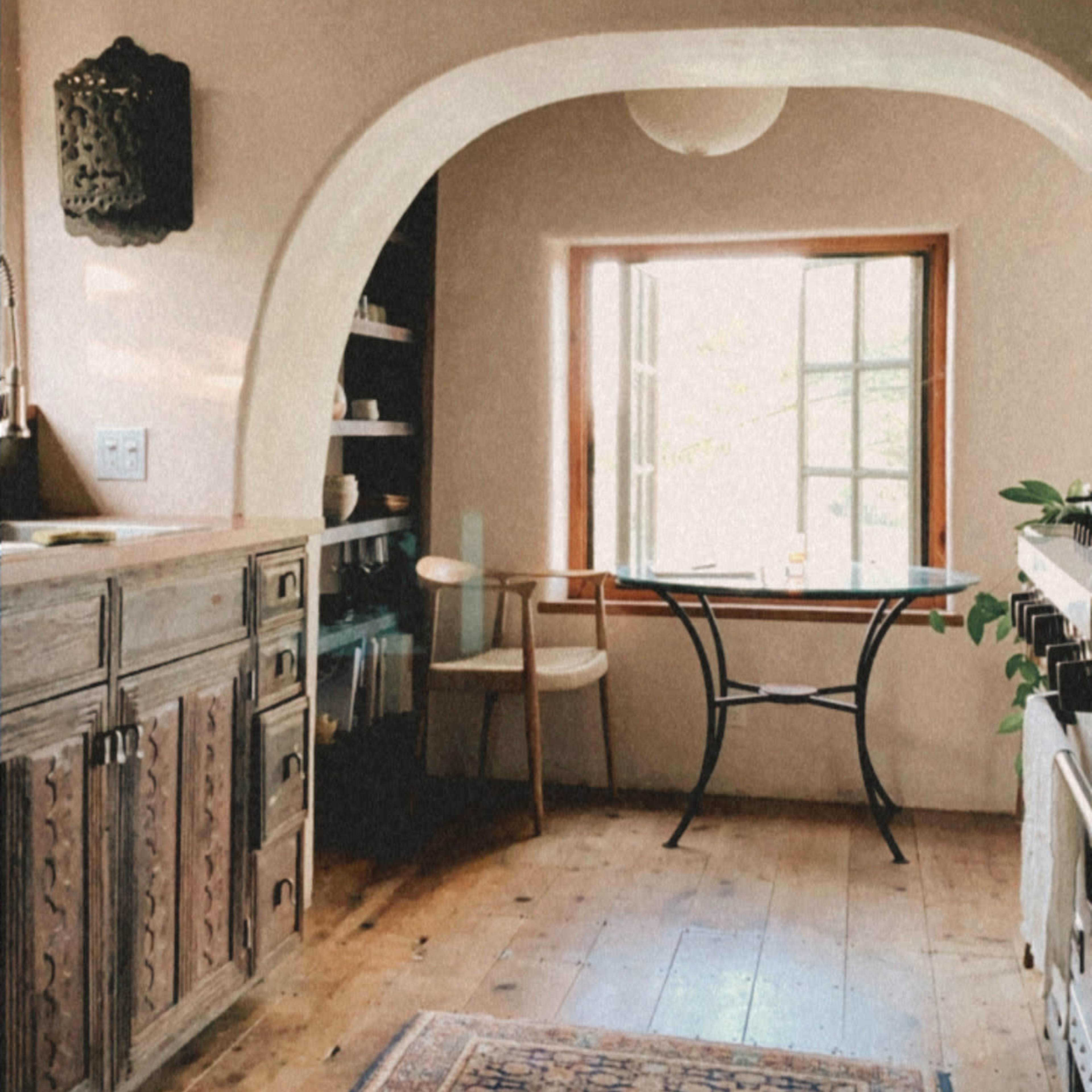 A rustic kitchen features wooden beams, a curved archway, a central table, and a window allowing natural light to enter.