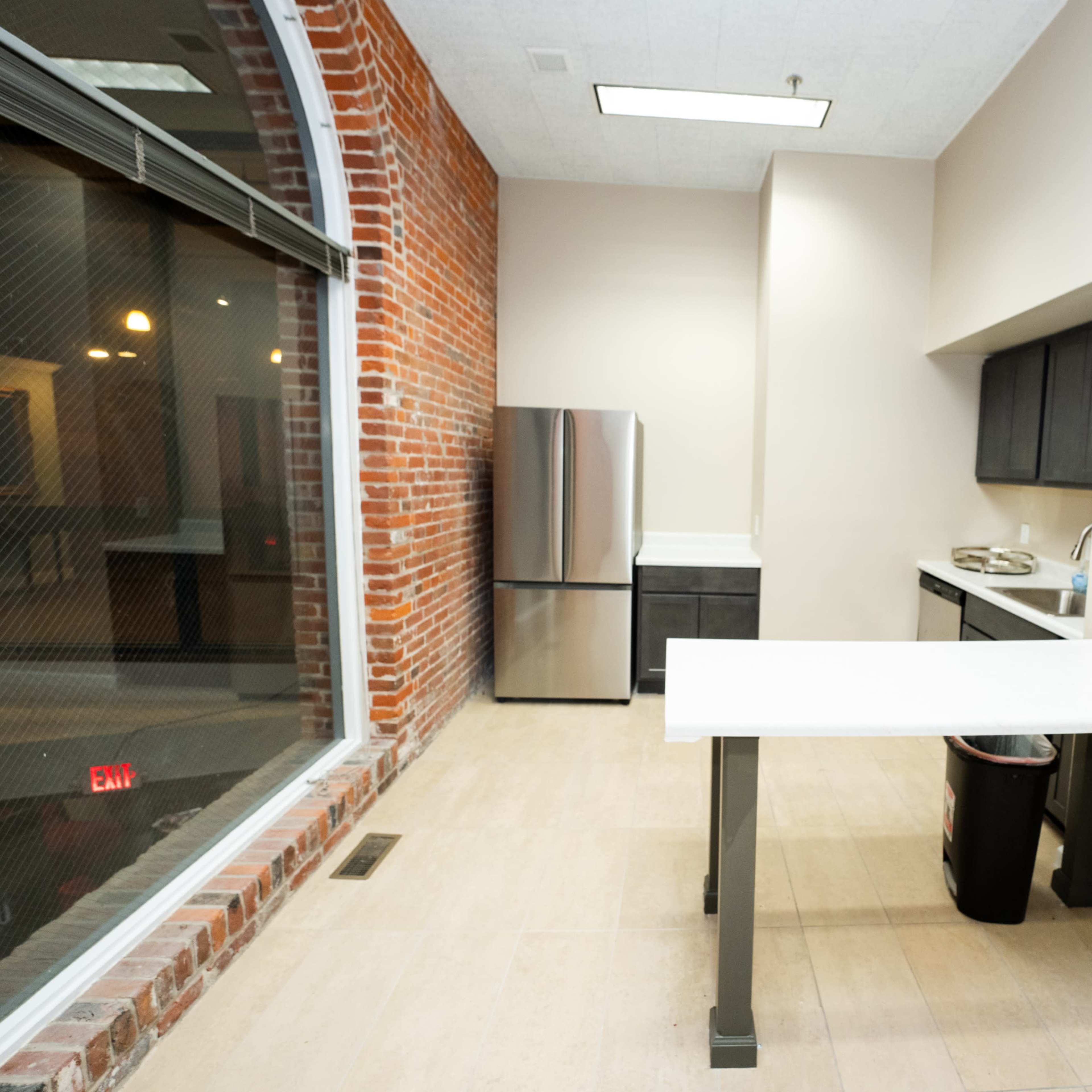A modern kitchen with a stainless steel refrigerator, black cabinets, and a white countertop table in a room with exposed brick walls.