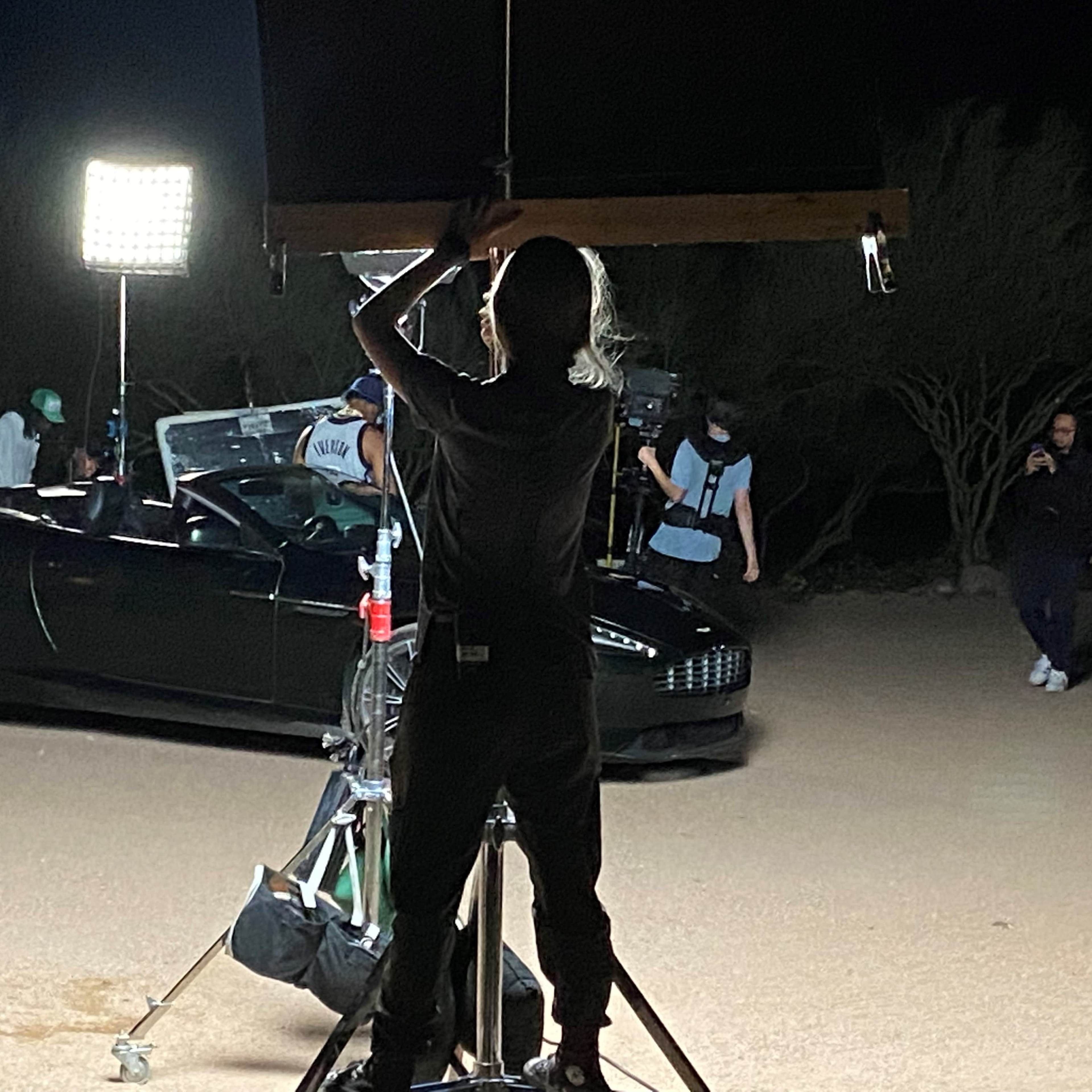 A crew member adjusts lighting equipment near a convertible car in a desert setting at night.
