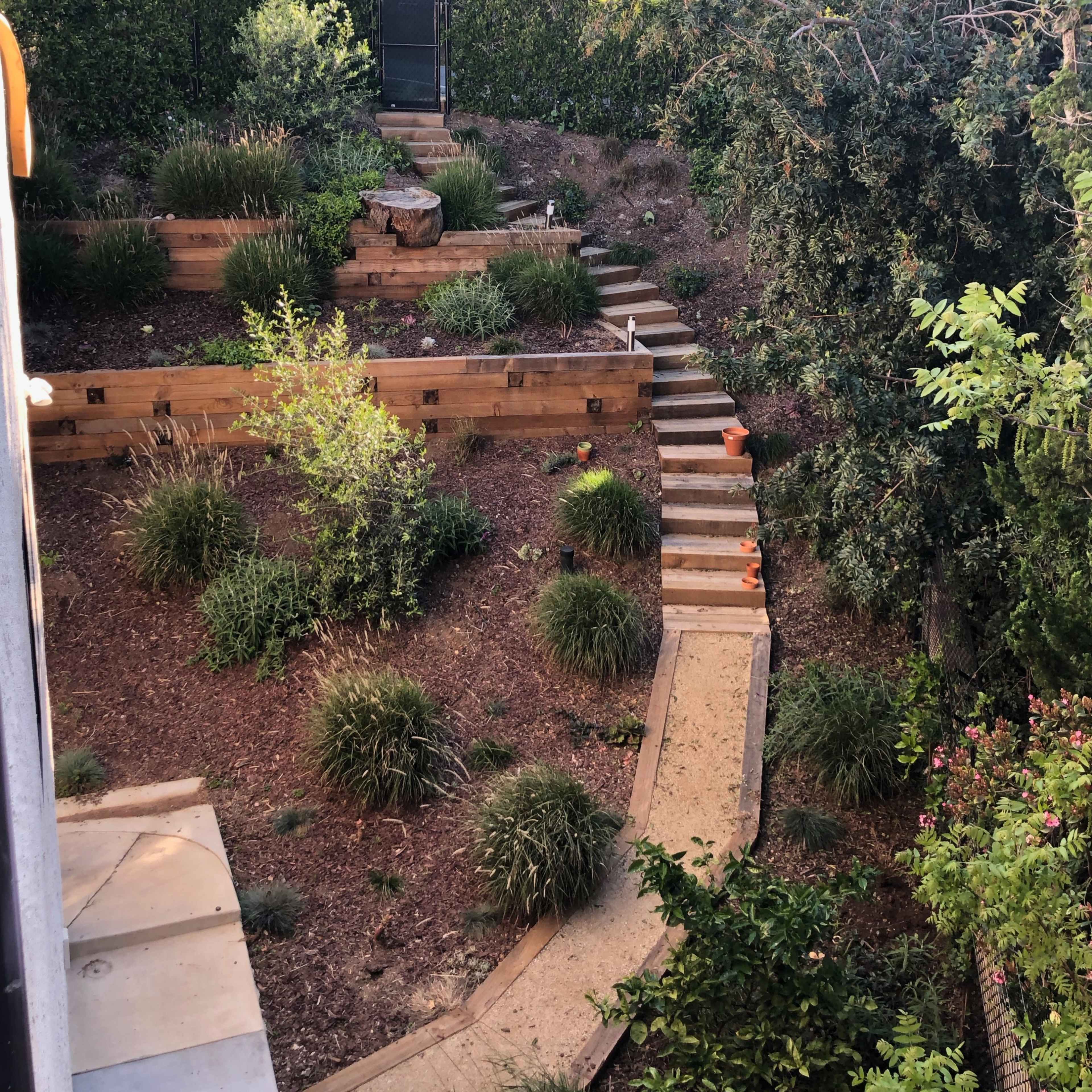 A landscaped garden features a series of wooden terraced beds, a pathway made of stone, and various plants set against a backdrop of dense greenery.