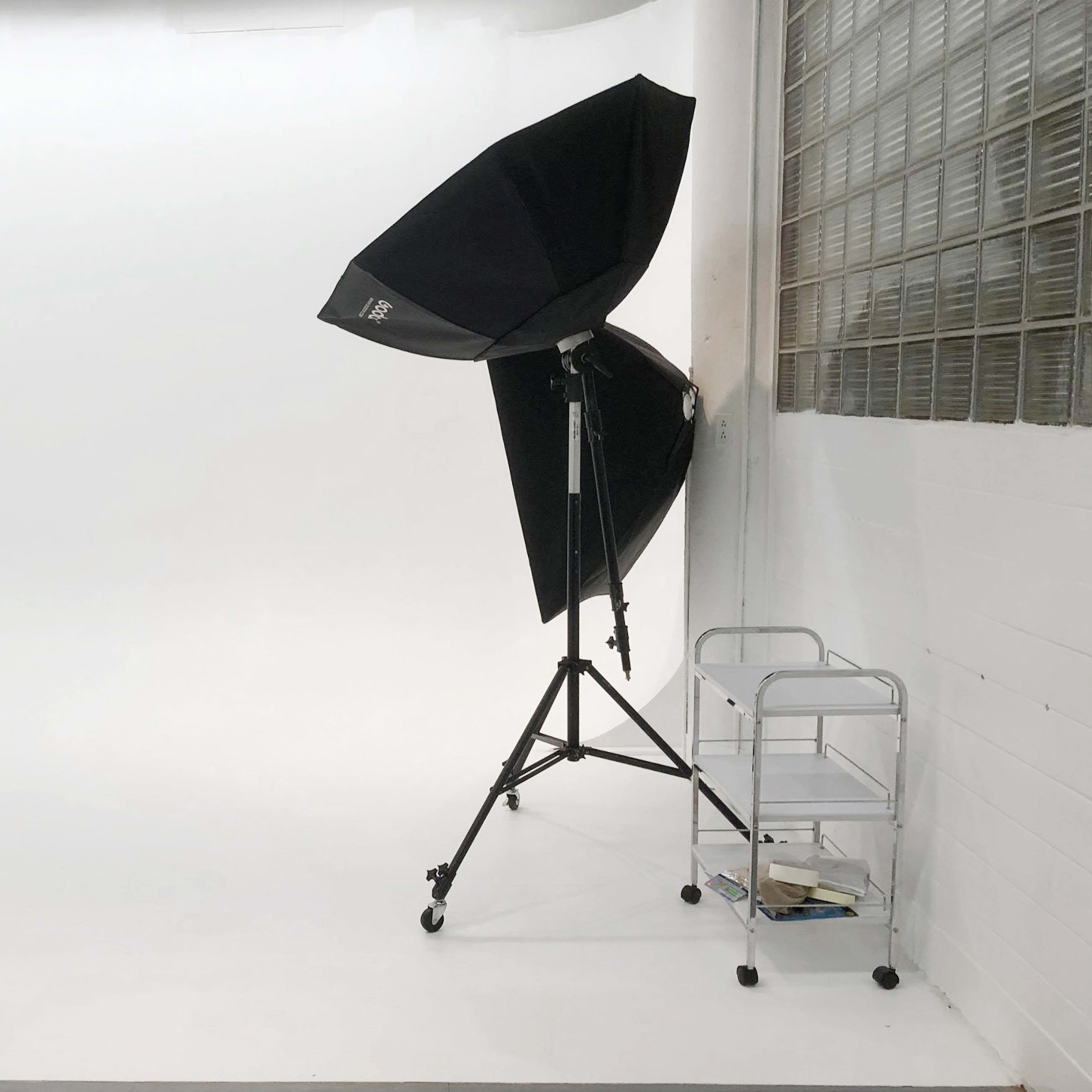 A photography studio features a smooth white backdrop, a large softbox umbrella light on a stand, and a small cart with some items beside it.