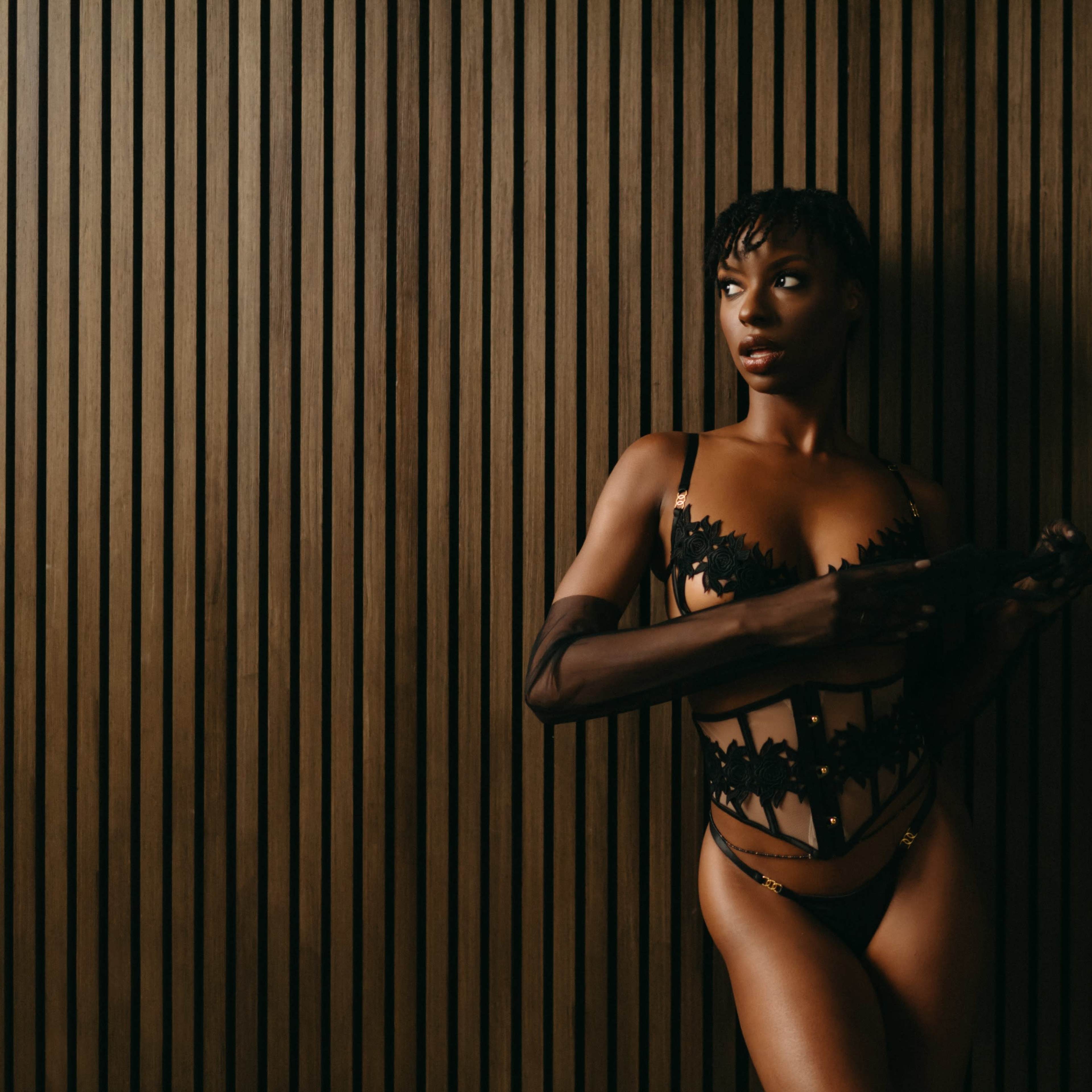 A model poses against a vertical wooden panel backdrop while wearing a black lingerie set.