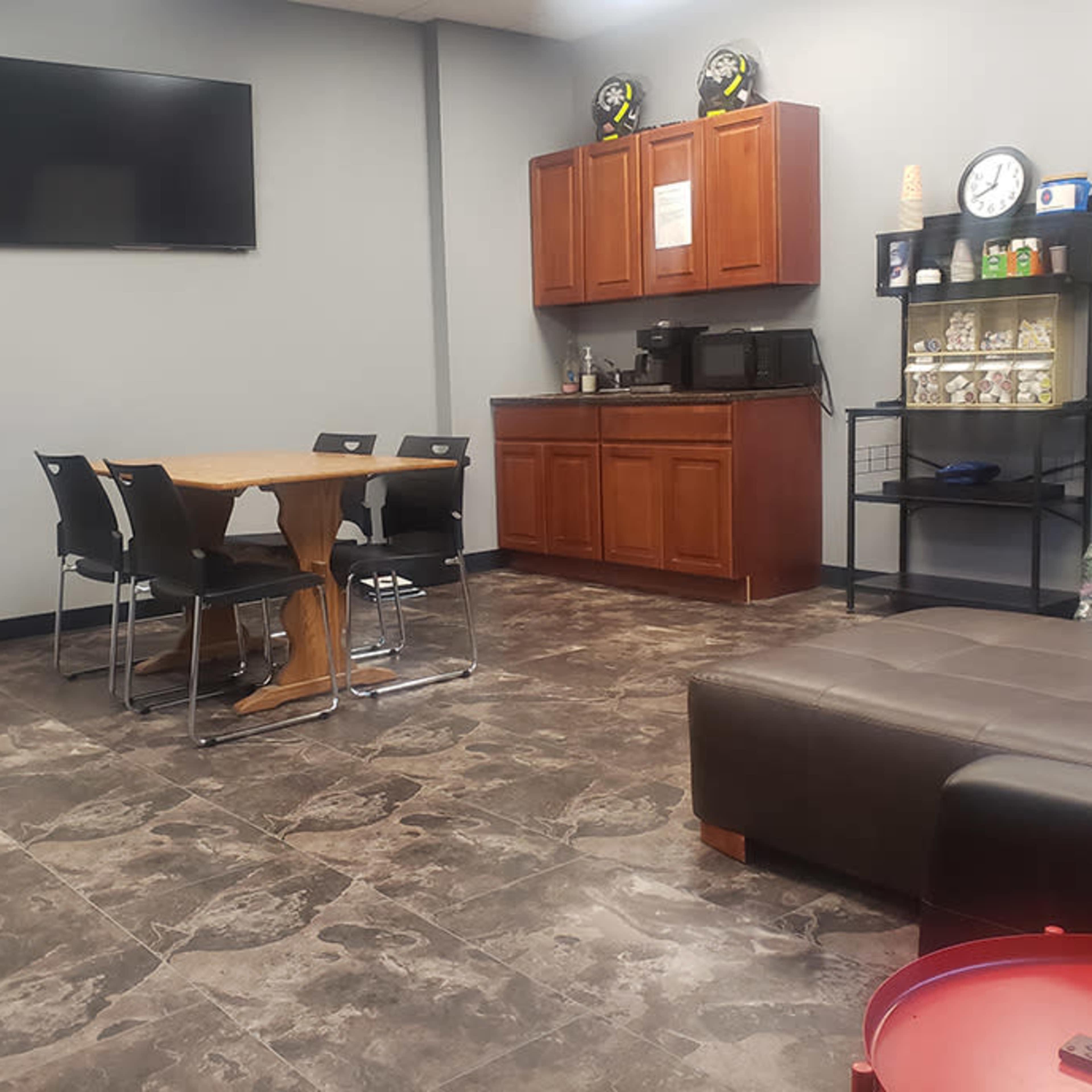 A break room with a wooden dining table and chairs, a kitchenette with cabinets, a microwave, and a television mounted on the wall.