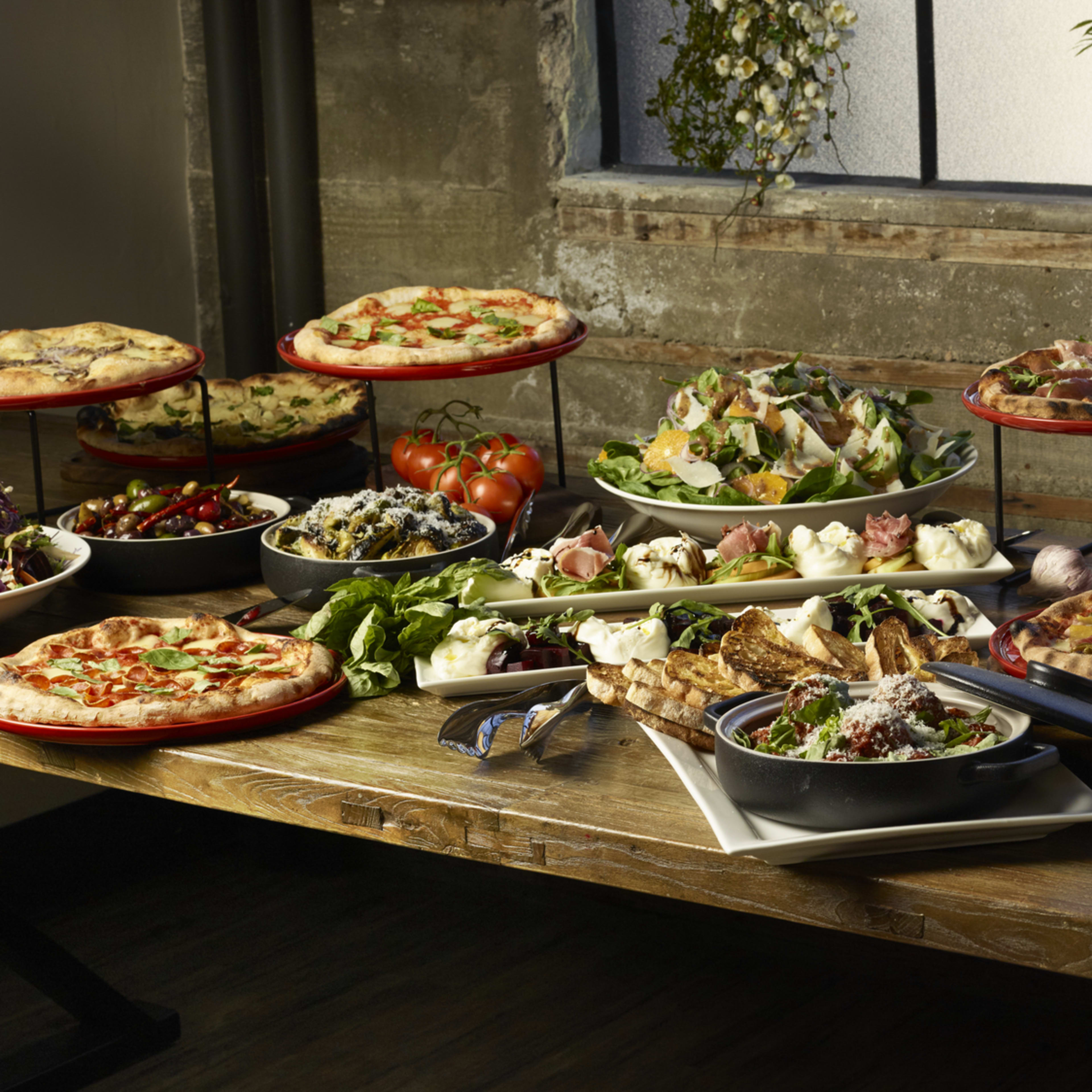 A long wooden table is set with various dishes, including pizzas, salads, and side items, surrounded by plants and natural light.