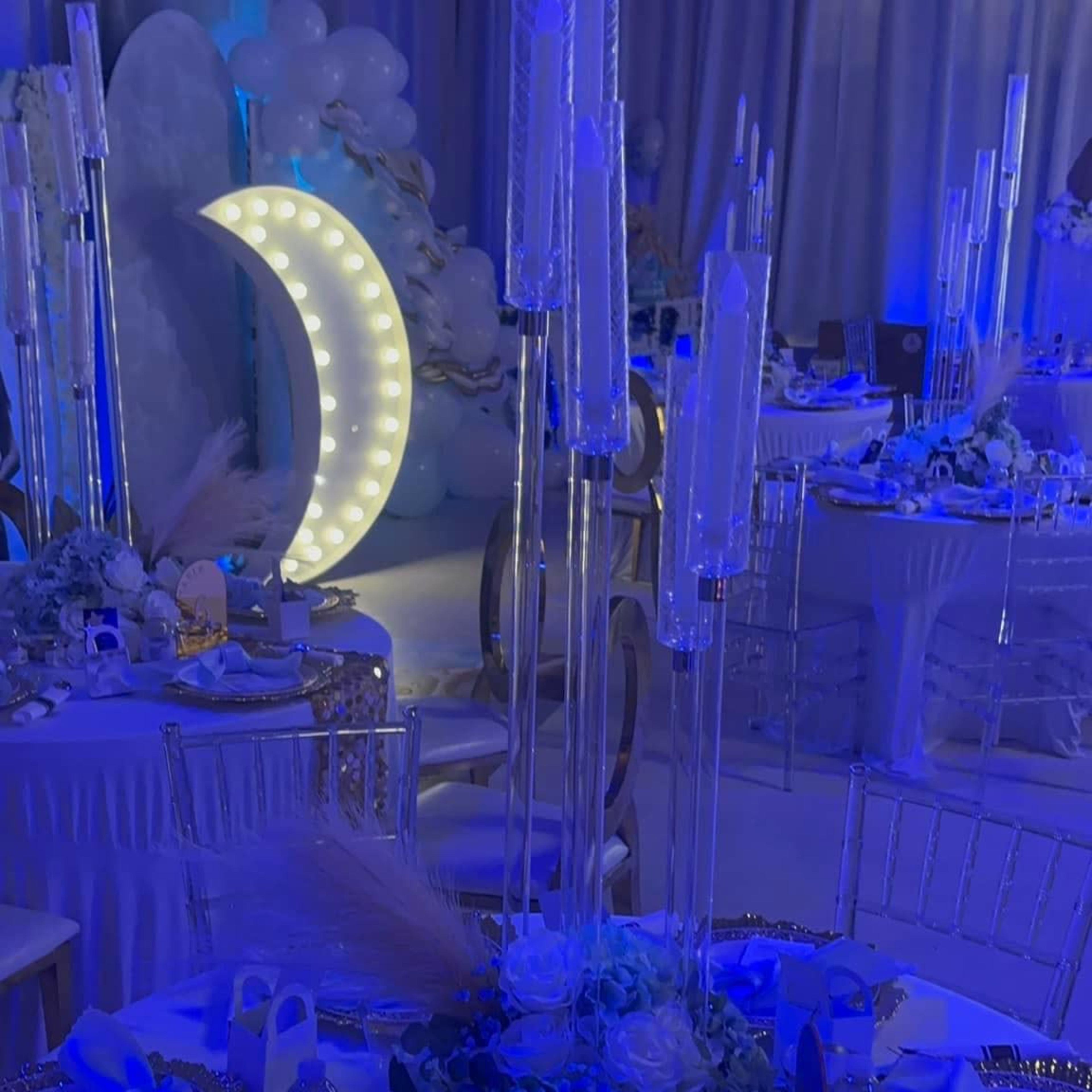 The table is elegantly set with blue lighting, featuring tall glass candle holders, floral arrangements, and gold-trimmed plates.