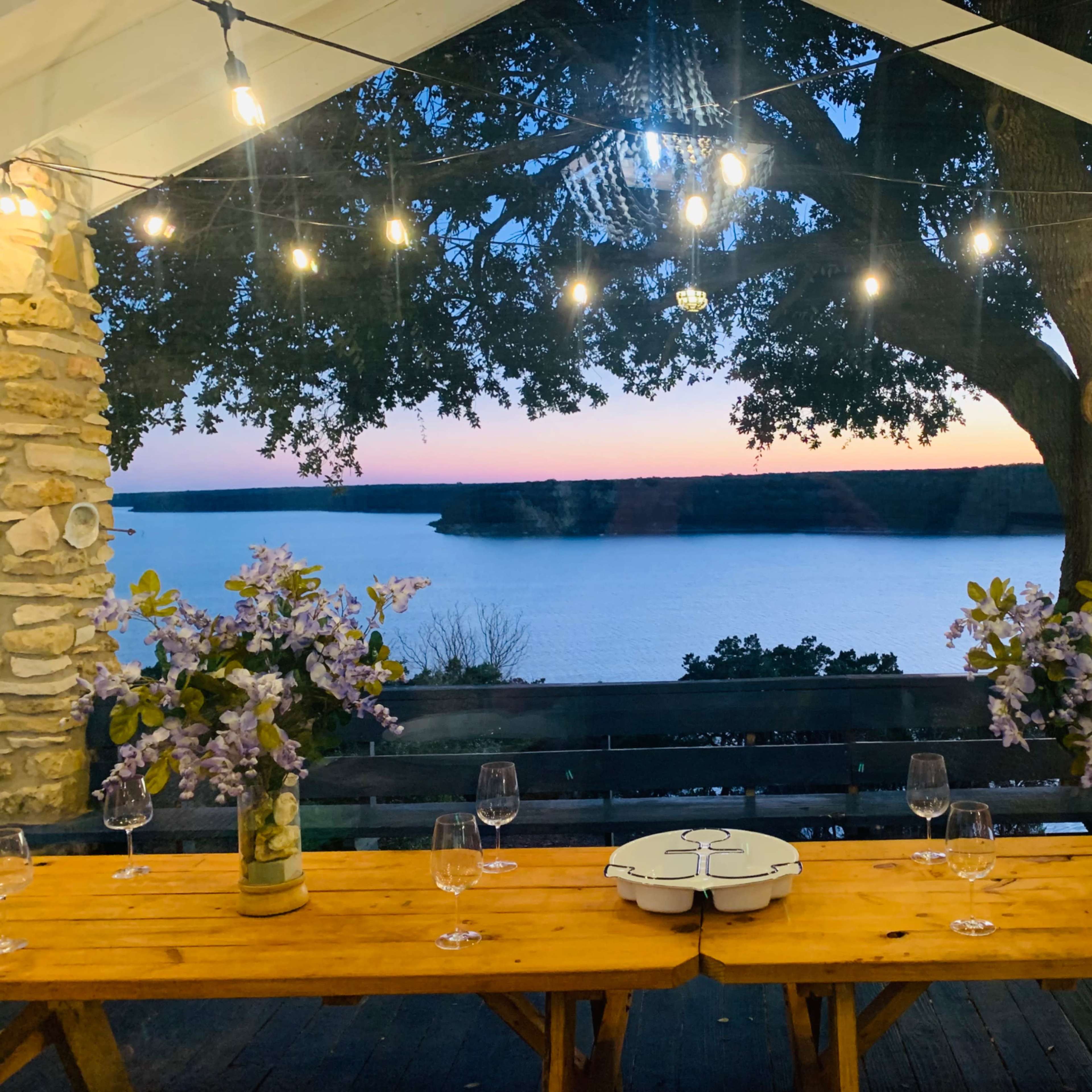 A rustic dining setup with wooden tables adorned with flower arrangements and wine glasses overlooks a calm lake at sunset, illuminated by string lights.