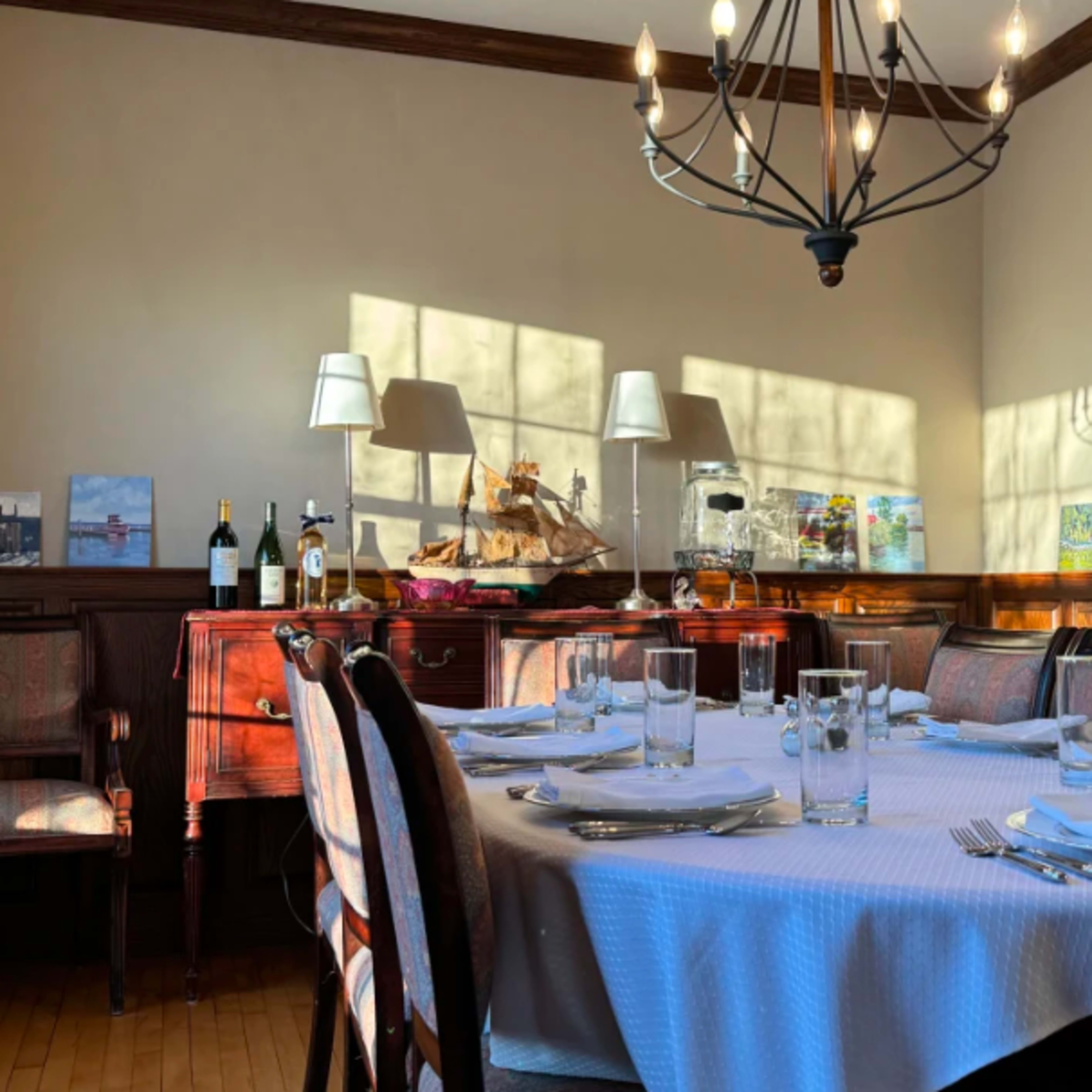 A dining room with a large round table set for a meal, surrounded by wooden chairs, and decorated with various artworks, wine bottles, and a chandelier overhead.