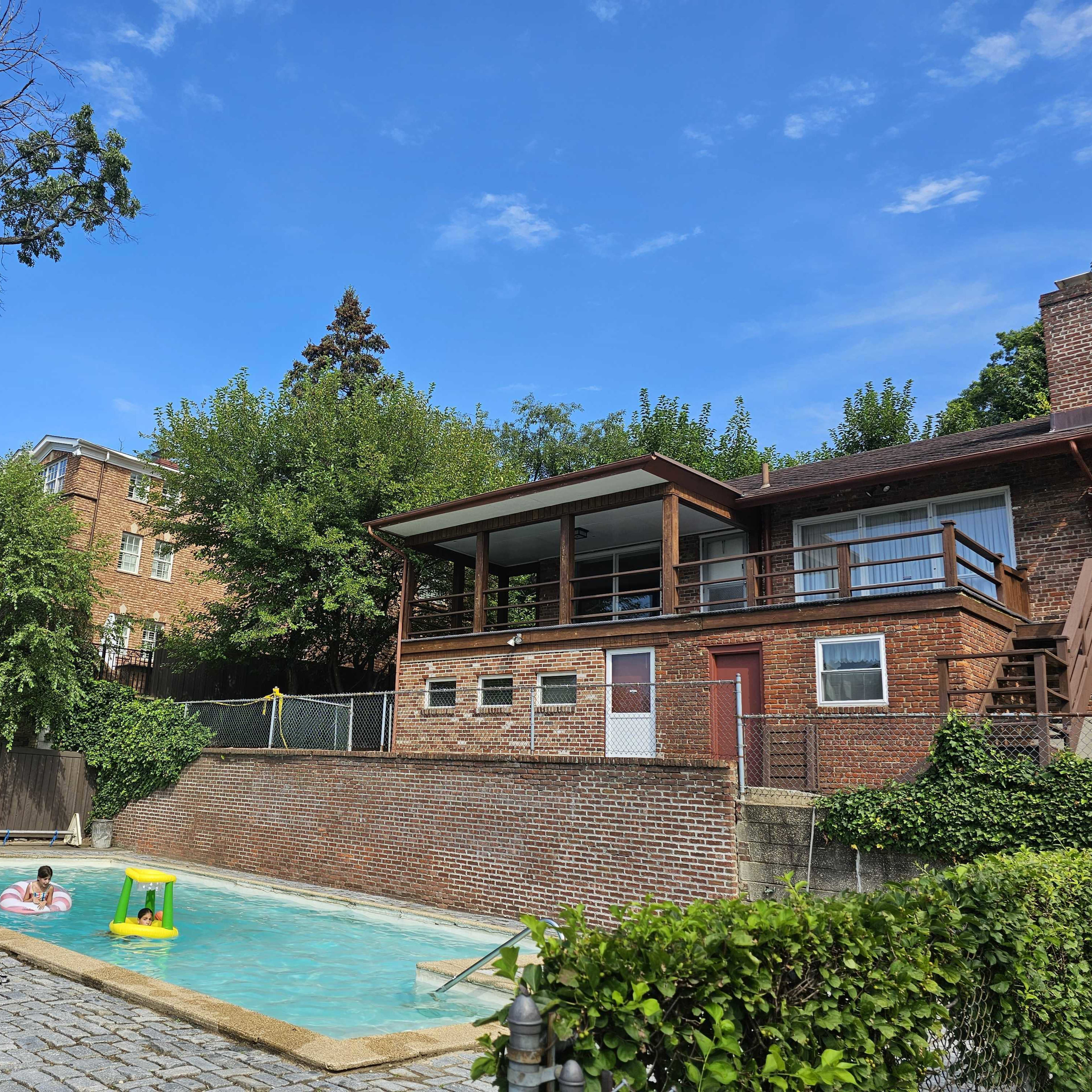 A backyard with a swimming pool surrounded by trees, a brick house with a balcony, and several people enjoying the water.