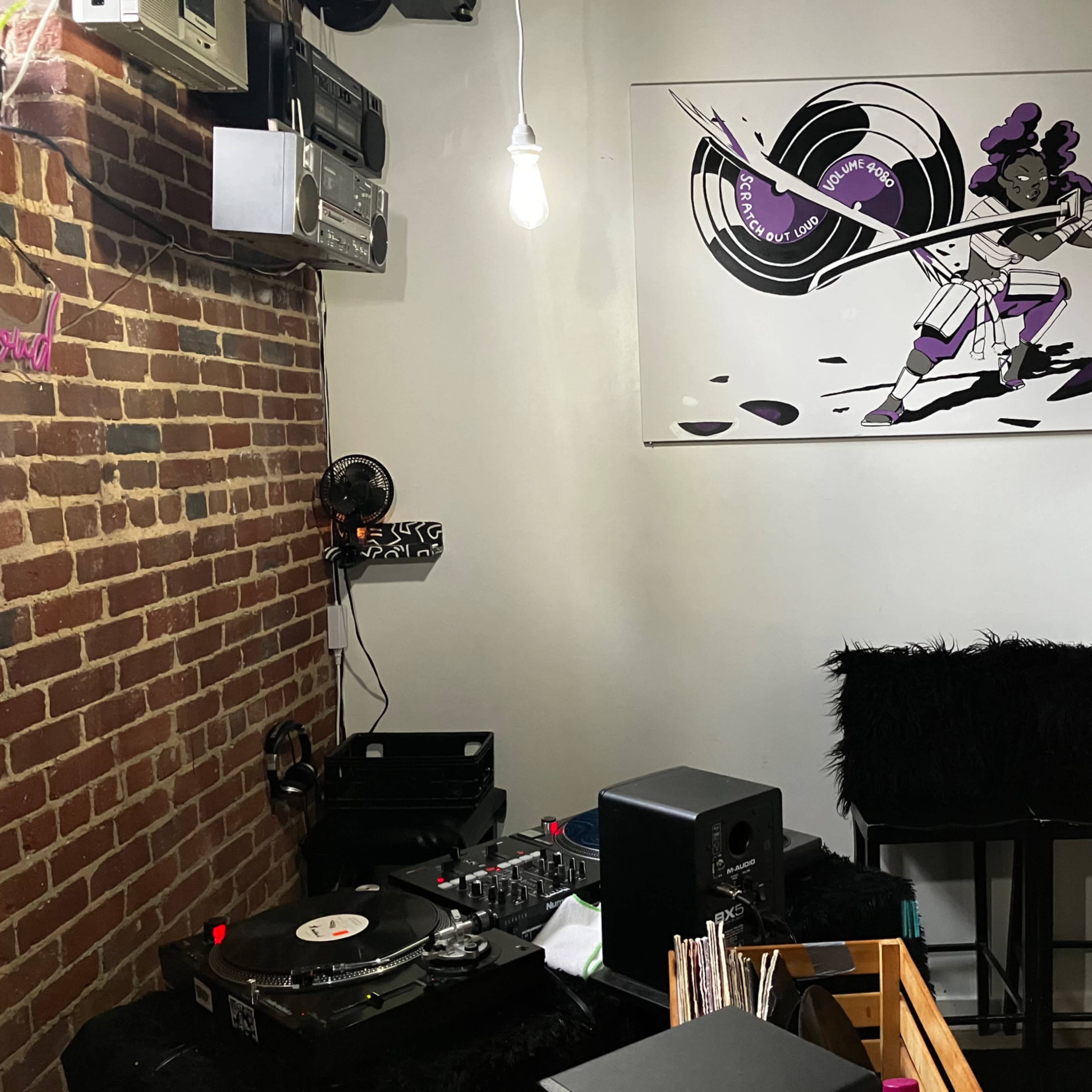 The room features a turntable setup on a black surface against a brick wall, with a large framed artwork of a musician playing an electric guitar above it.