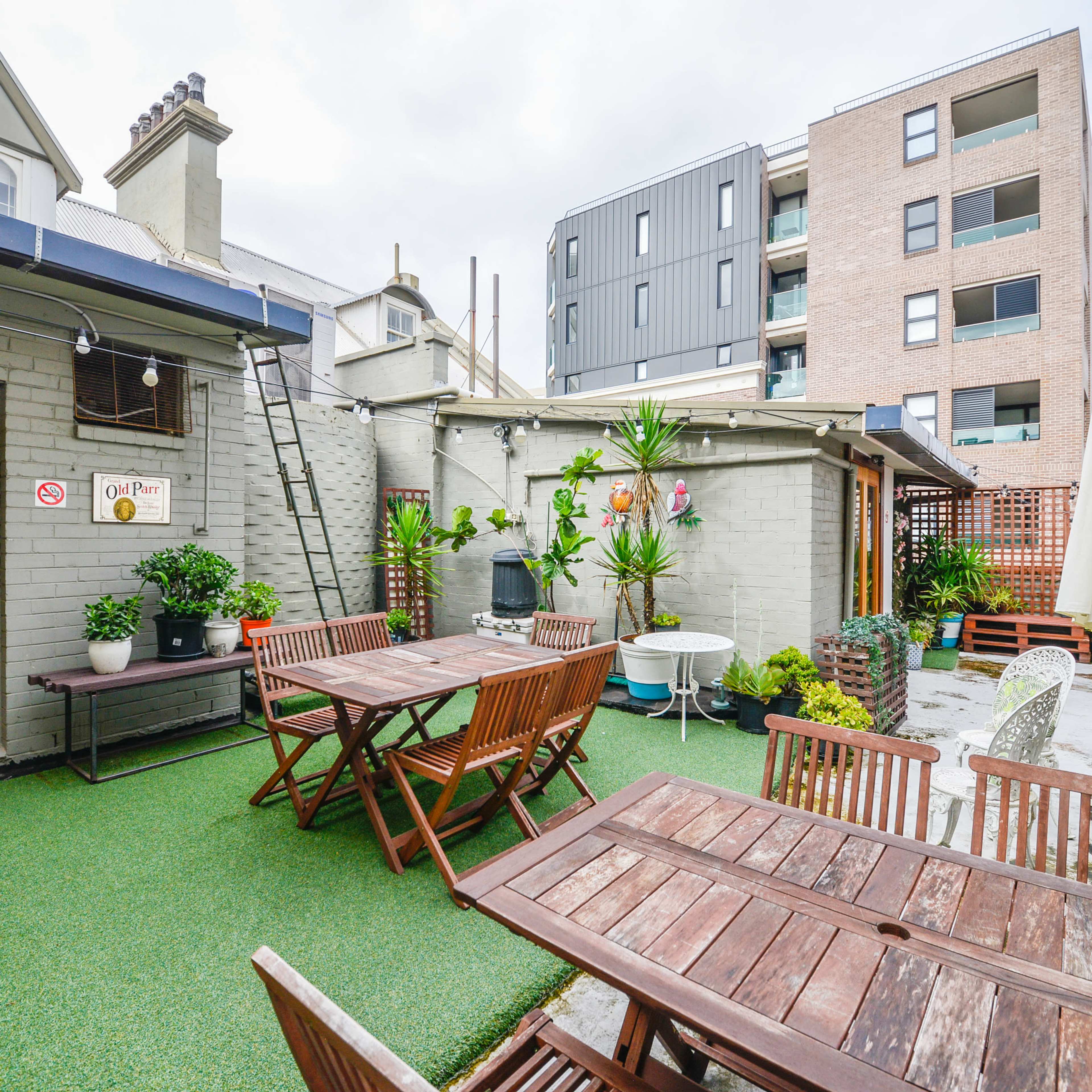 A patio area with wooden tables and chairs, surrounded by potted plants, against a backdrop of both a traditional and modern building.