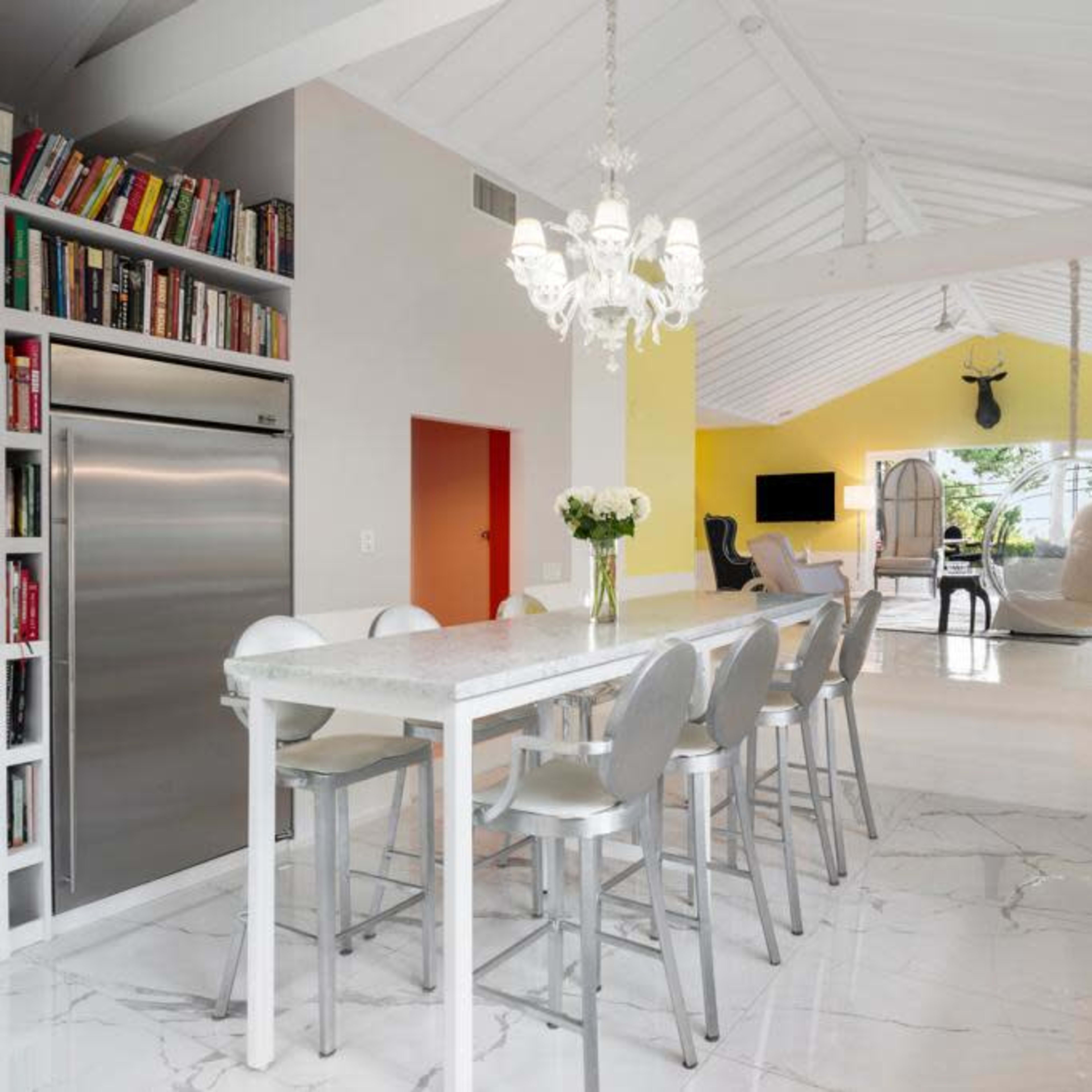A modern kitchen features a long white dining table with high chairs, large windows, and an open layout leading to a bright yellow living area.