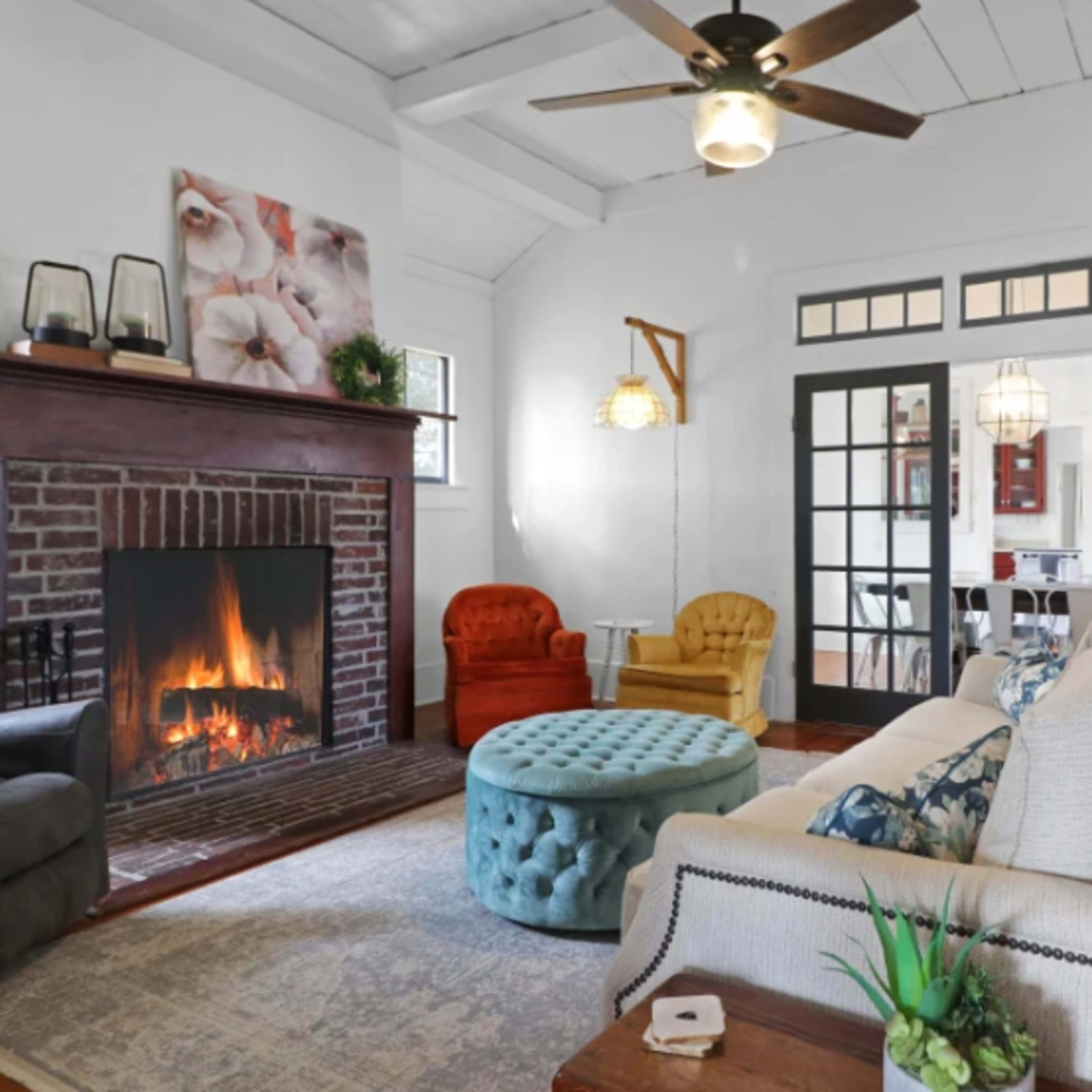 The living room features a fireplace, comfortable seating, and a blue ottoman, with large windows allowing natural light to enter.
