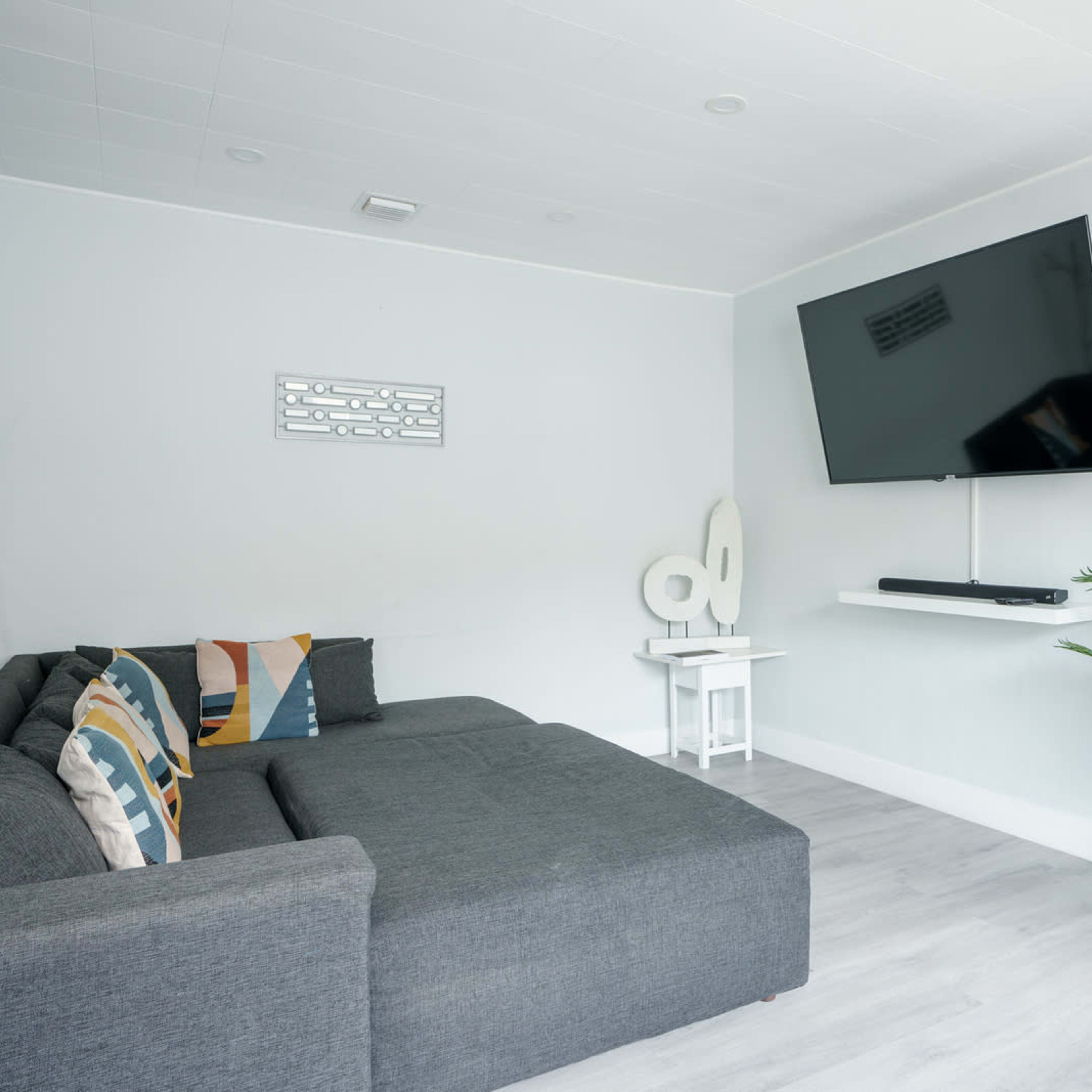 A modern living room features a gray sectional sofa, a mounted flat-screen TV, and a small indoor plant beside a white side table.