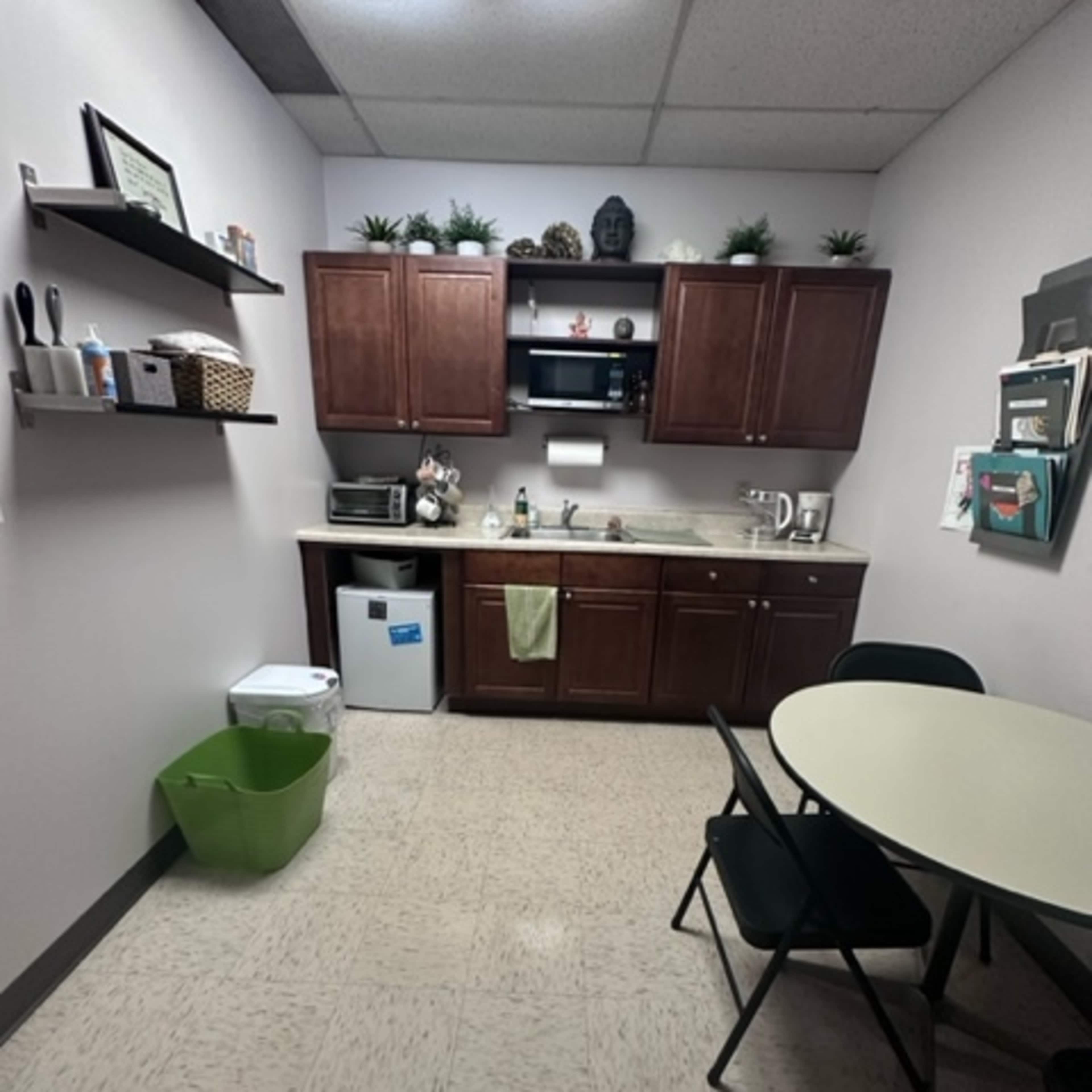 A small kitchen area features wooden cabinets, a microwave, a sink, and a table with chairs, along with a green trash bin in the corner.