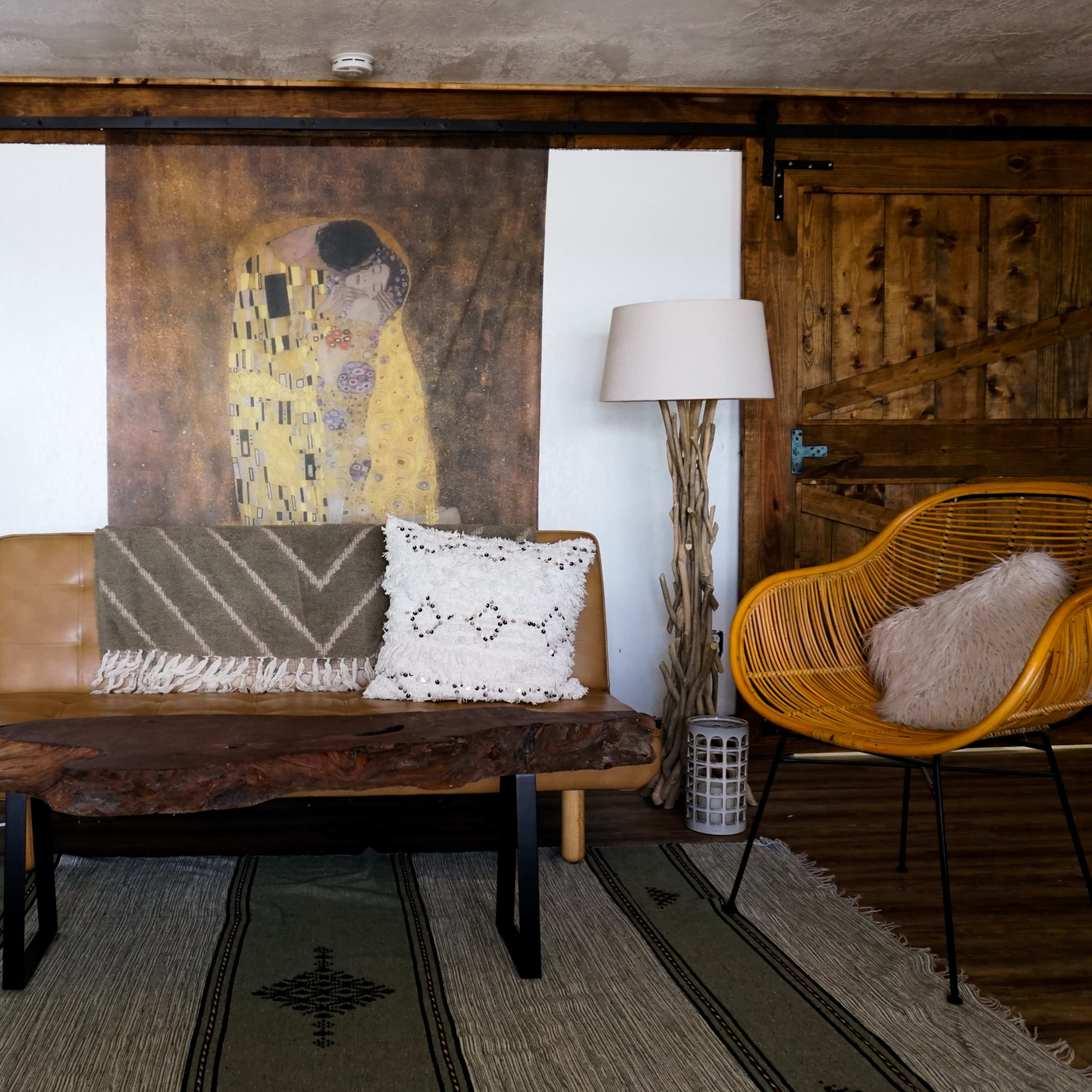 The scene features a living room with a leather sofa, two woven chairs, a large decorative wall art piece, and a patterned rug.