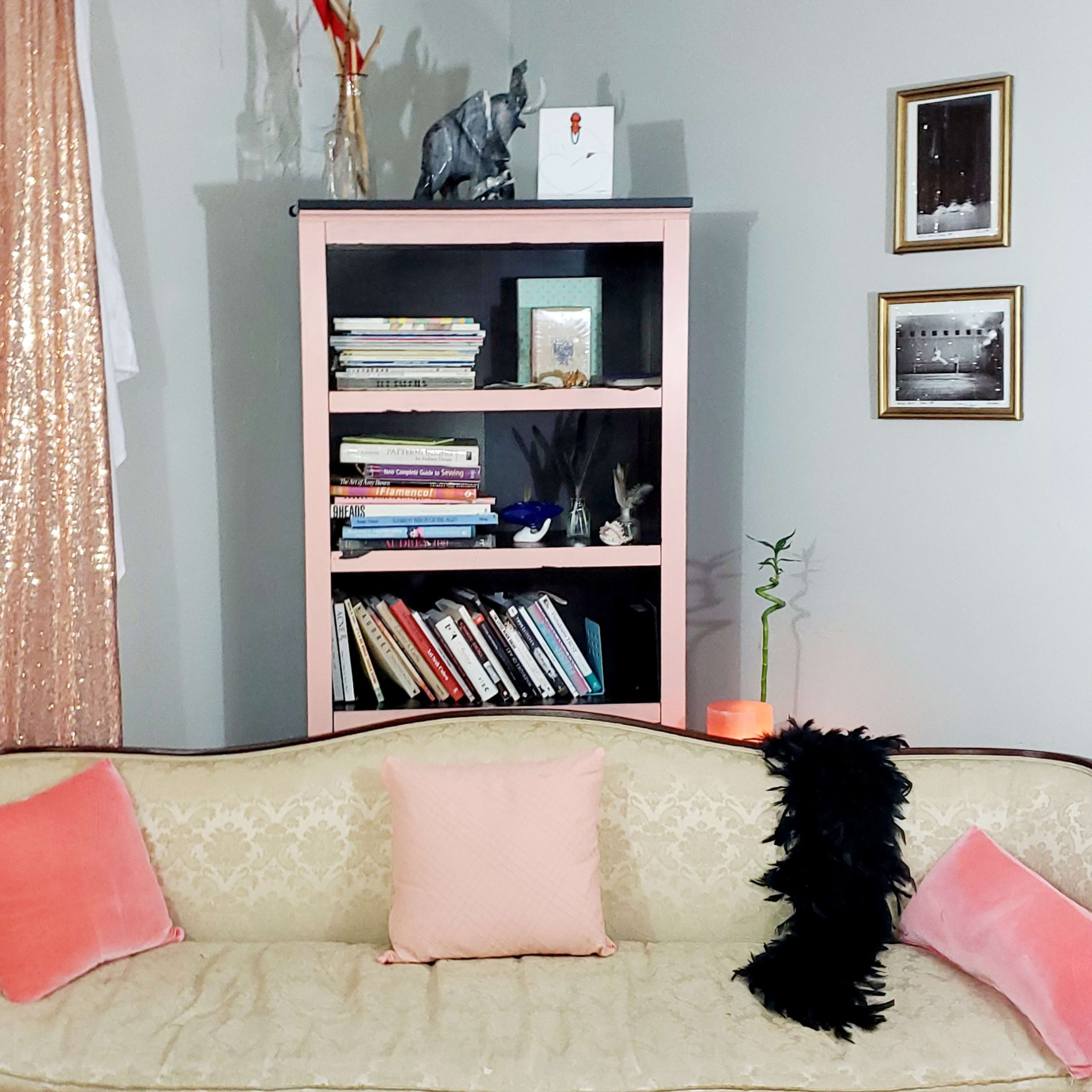 A living room with a vintage couch adorned with pink cushions, a bookshelf filled with books, and curtains that shimmer in a golden hue.