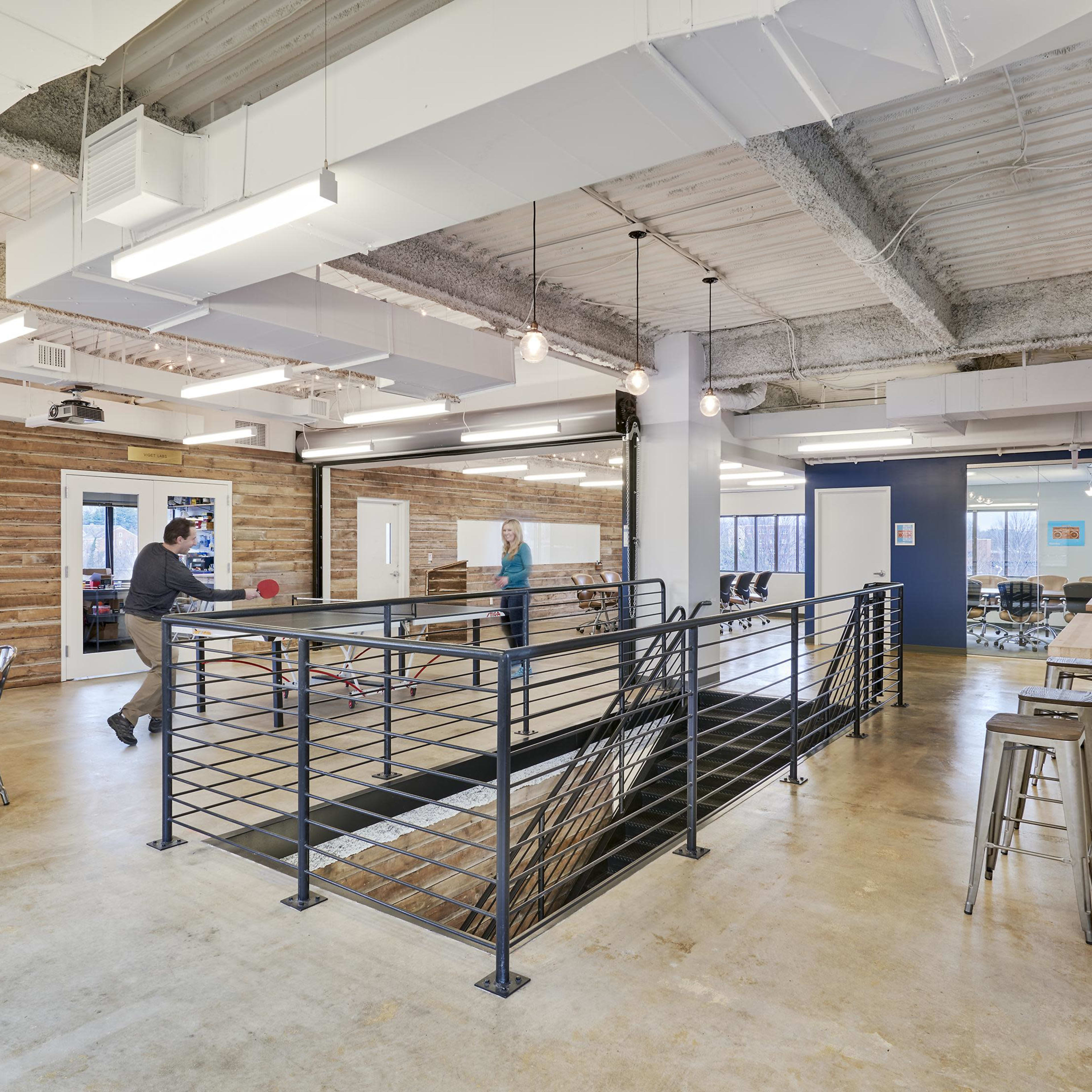 A modern open office space features a ping pong table, wooden tables, and a staircase leading to a lower level, with varied seating arrangements and industrial lighting.