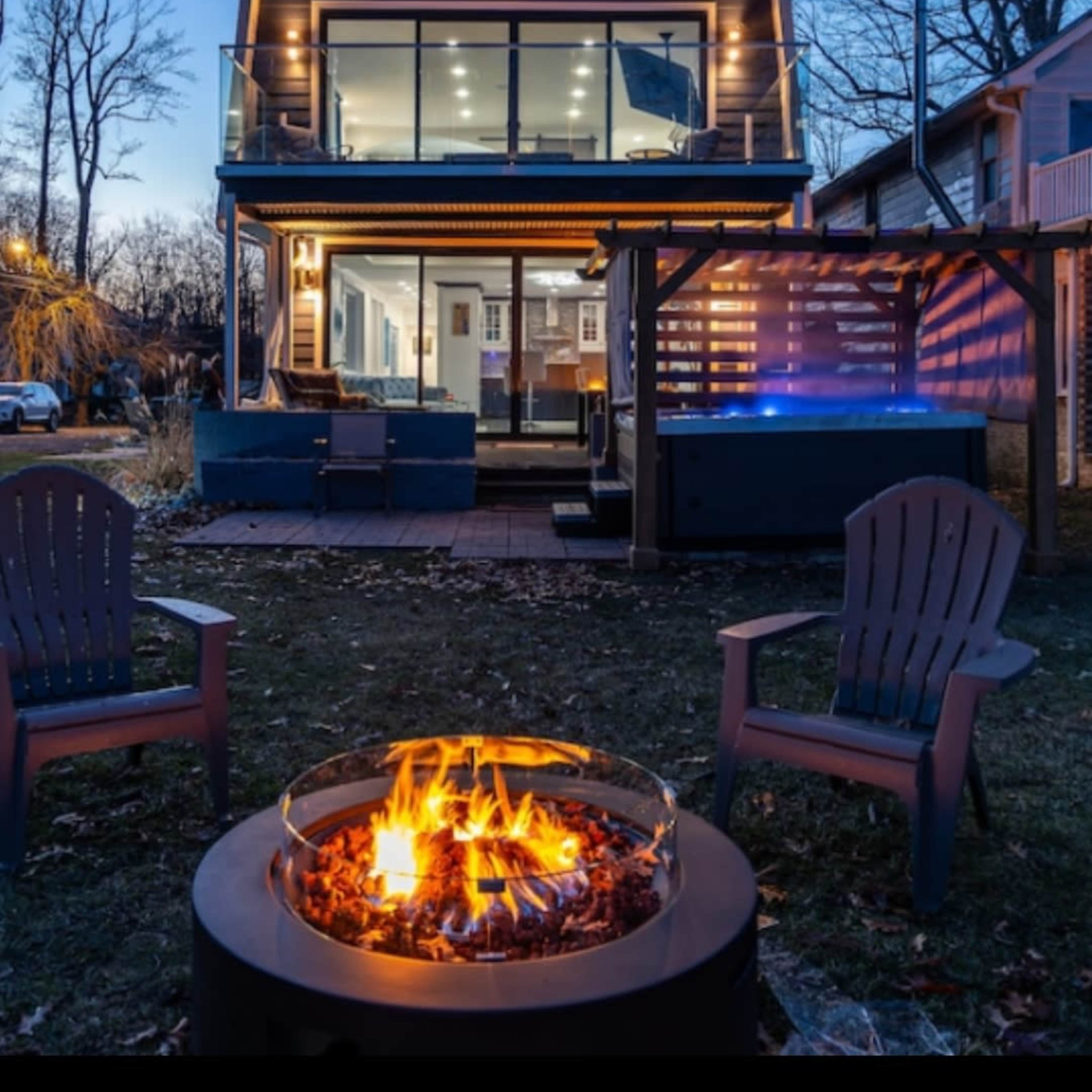 A modern two-story house with large windows and a deck overlooks a fire pit surrounded by wooden chairs, set against a twilight sky.