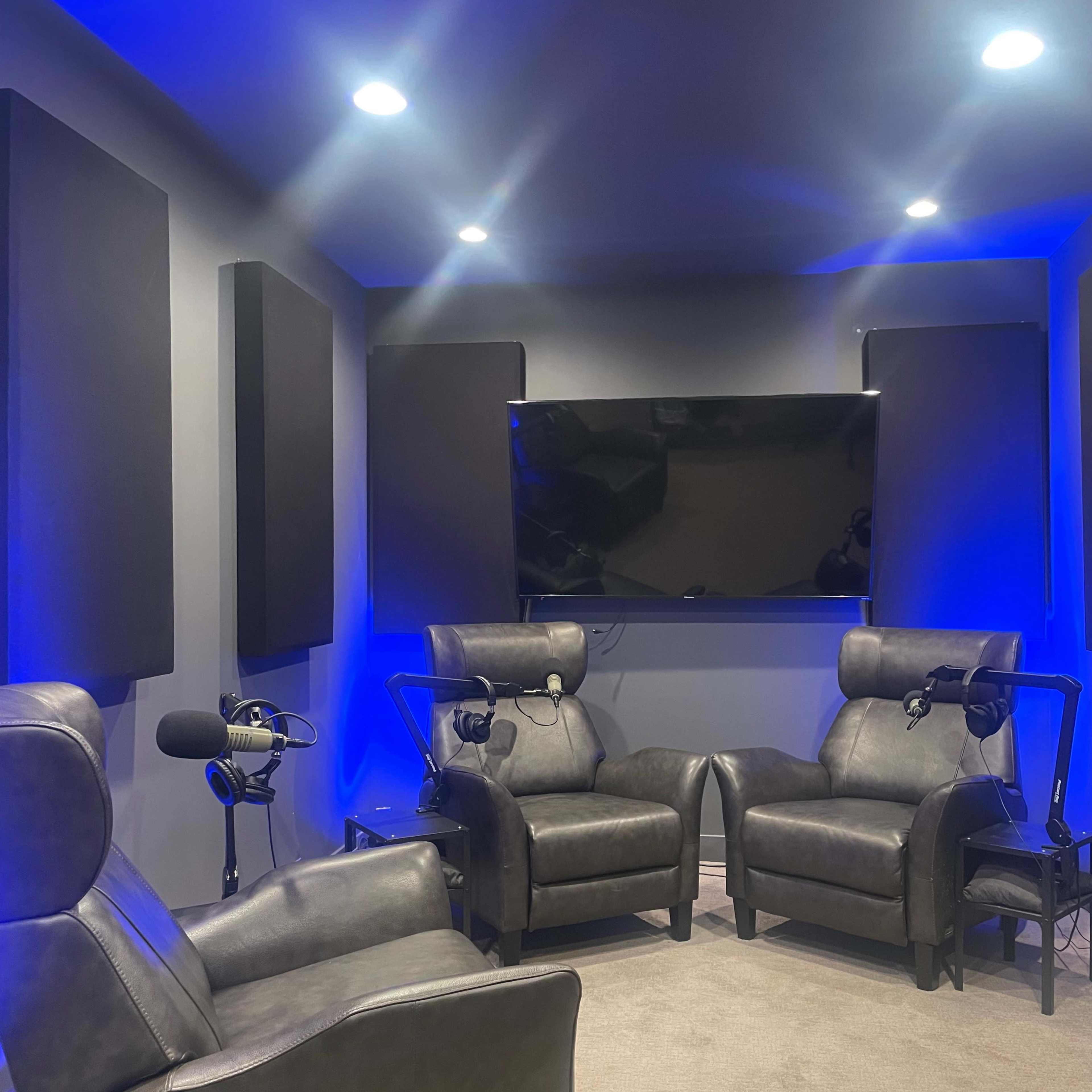A small recording studio featuring two leather chairs, a television mounted on the wall, and soundproof panels on the sides, illuminated by blue lighting.