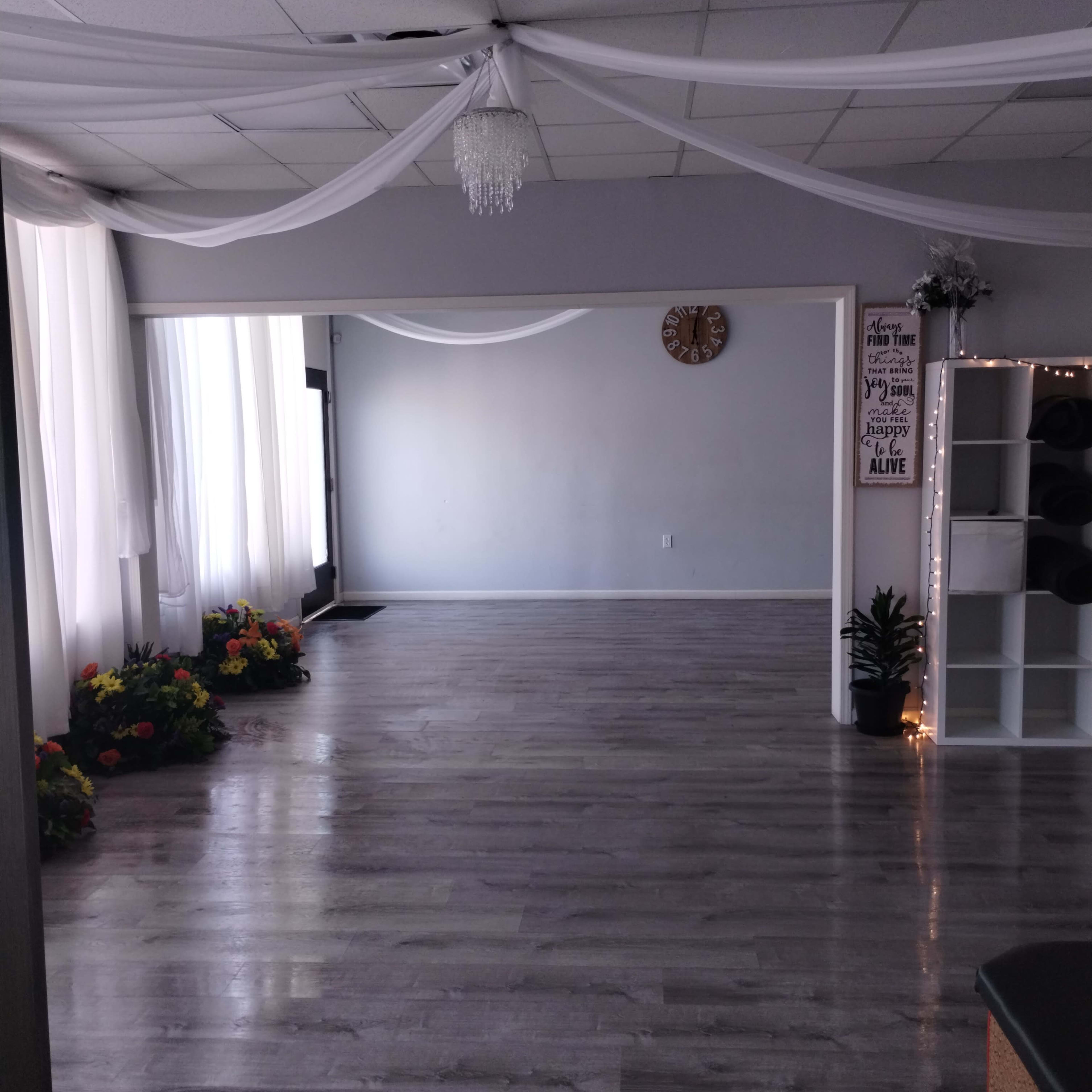A spacious studio with wooden flooring, large windows draped with white fabric, and flower arrangements in the corners.