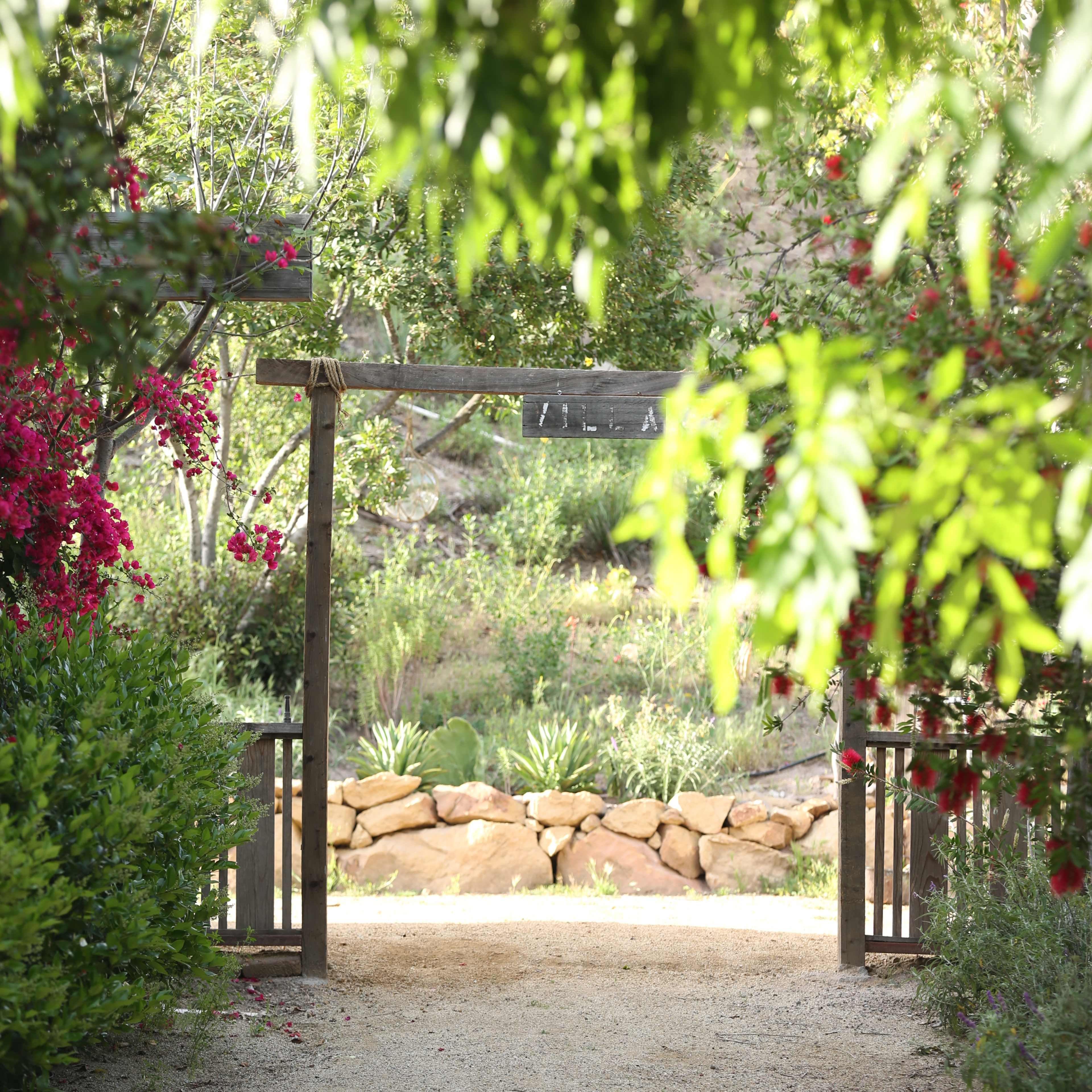 A pathway leads through a wooden archway into a lush garden with greenery and flowering plants.