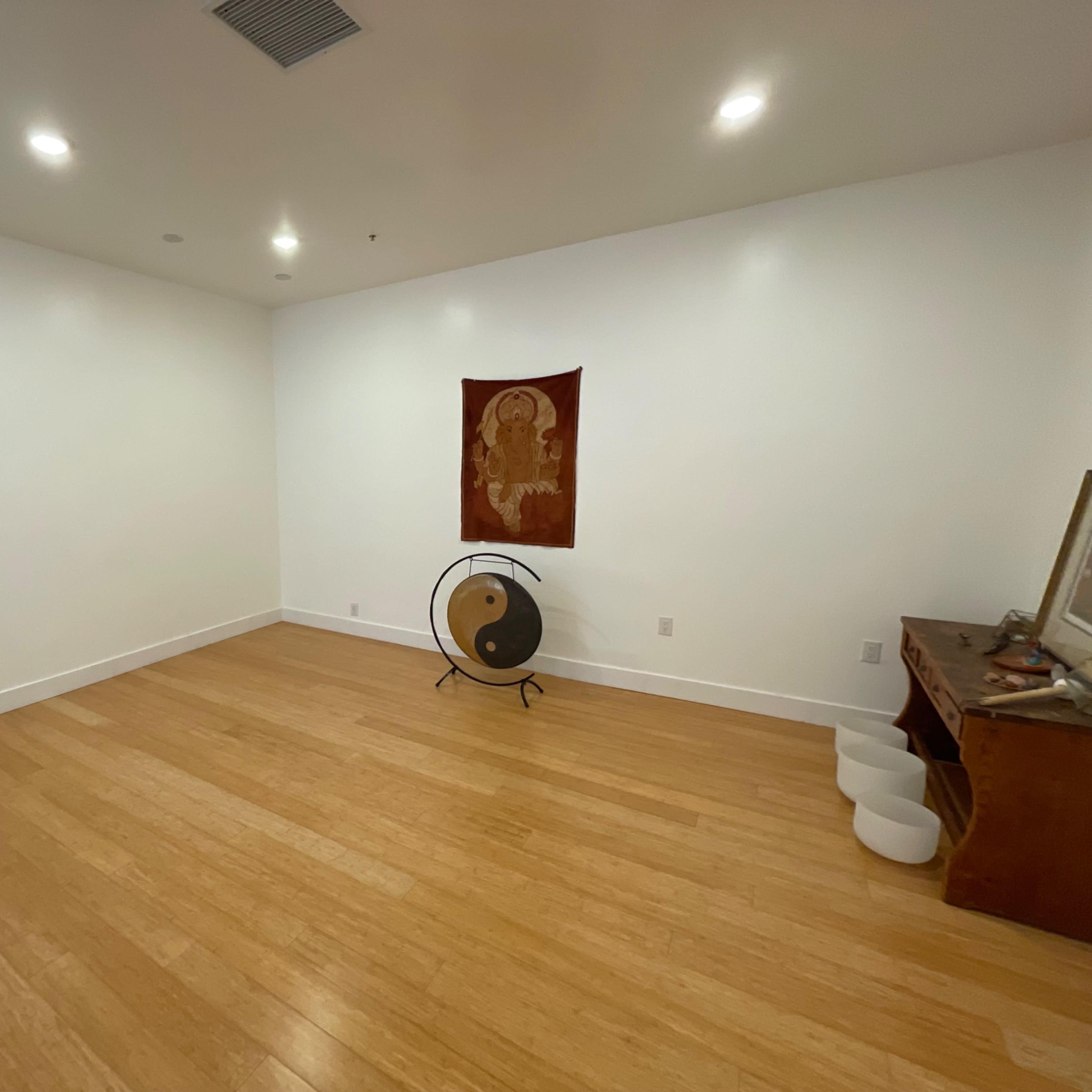 A sparsely furnished room with bamboo flooring, featuring a wall tapestry, a gong on a stand, and a wooden cabinet.