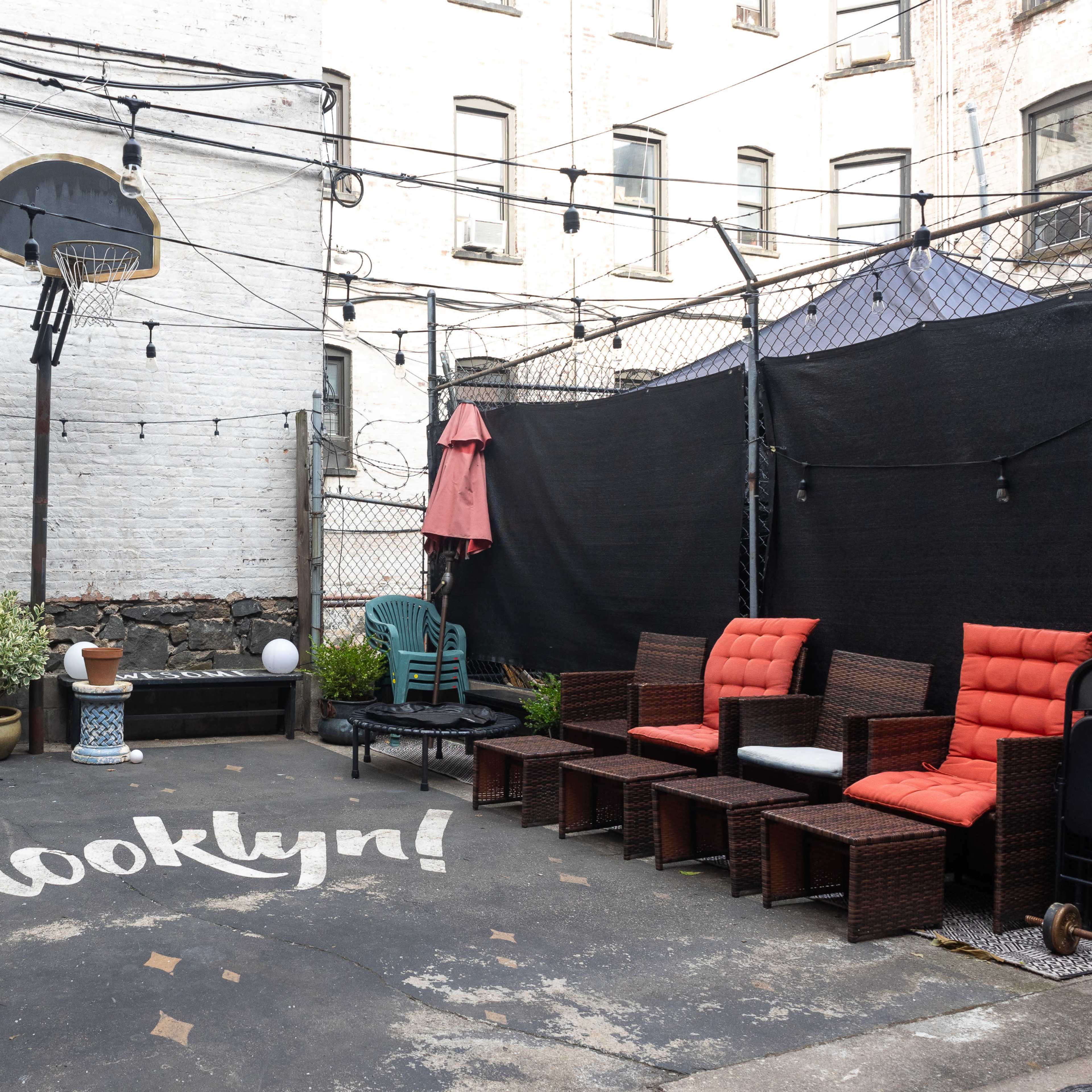A courtyard area in Brooklyn with outdoor seating, basketball hoop, string lights, and a painted "Brooklyn" on the ground.