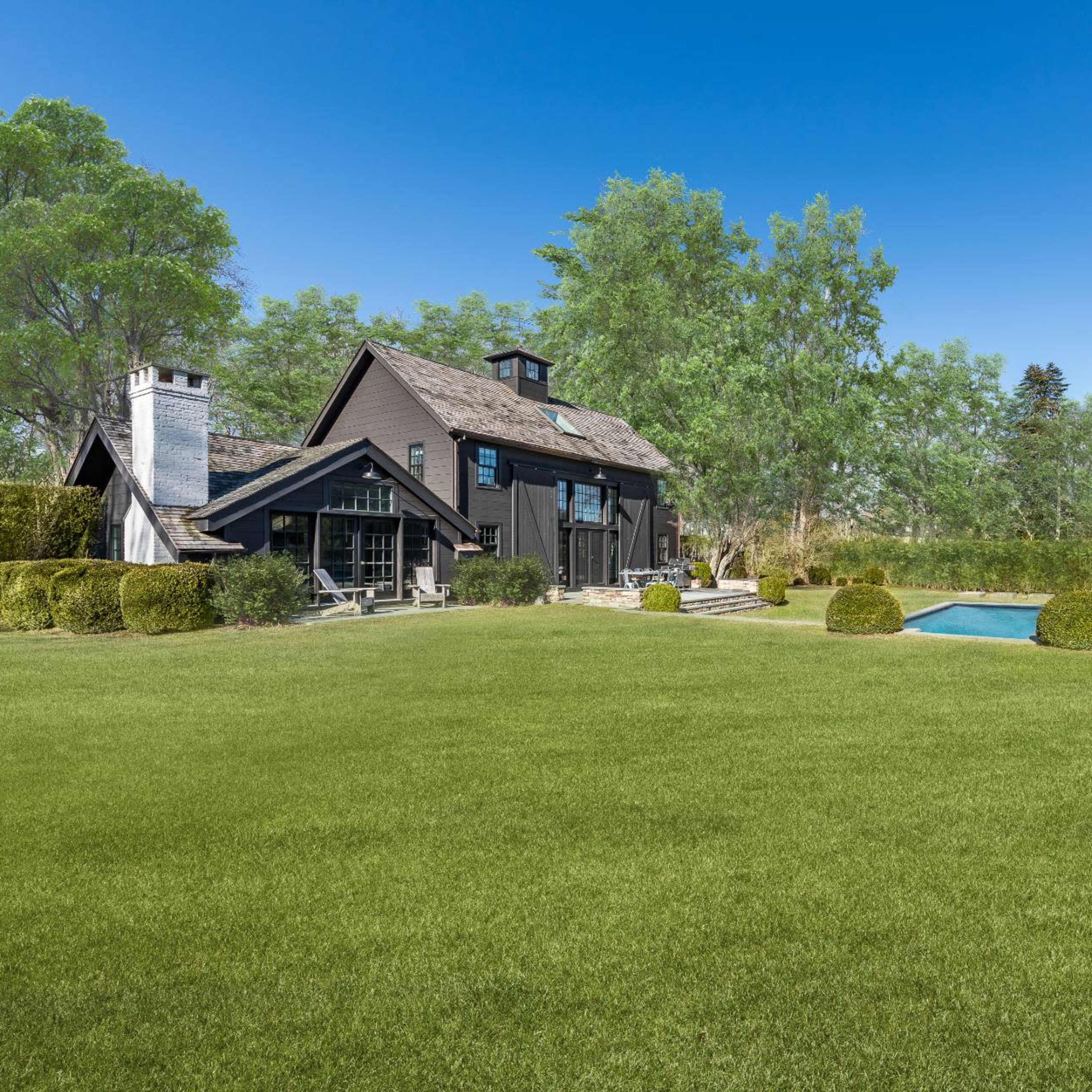 A black wooden house with a chimney, surrounded by a well-manicured lawn and a swimming pool in the backyard.