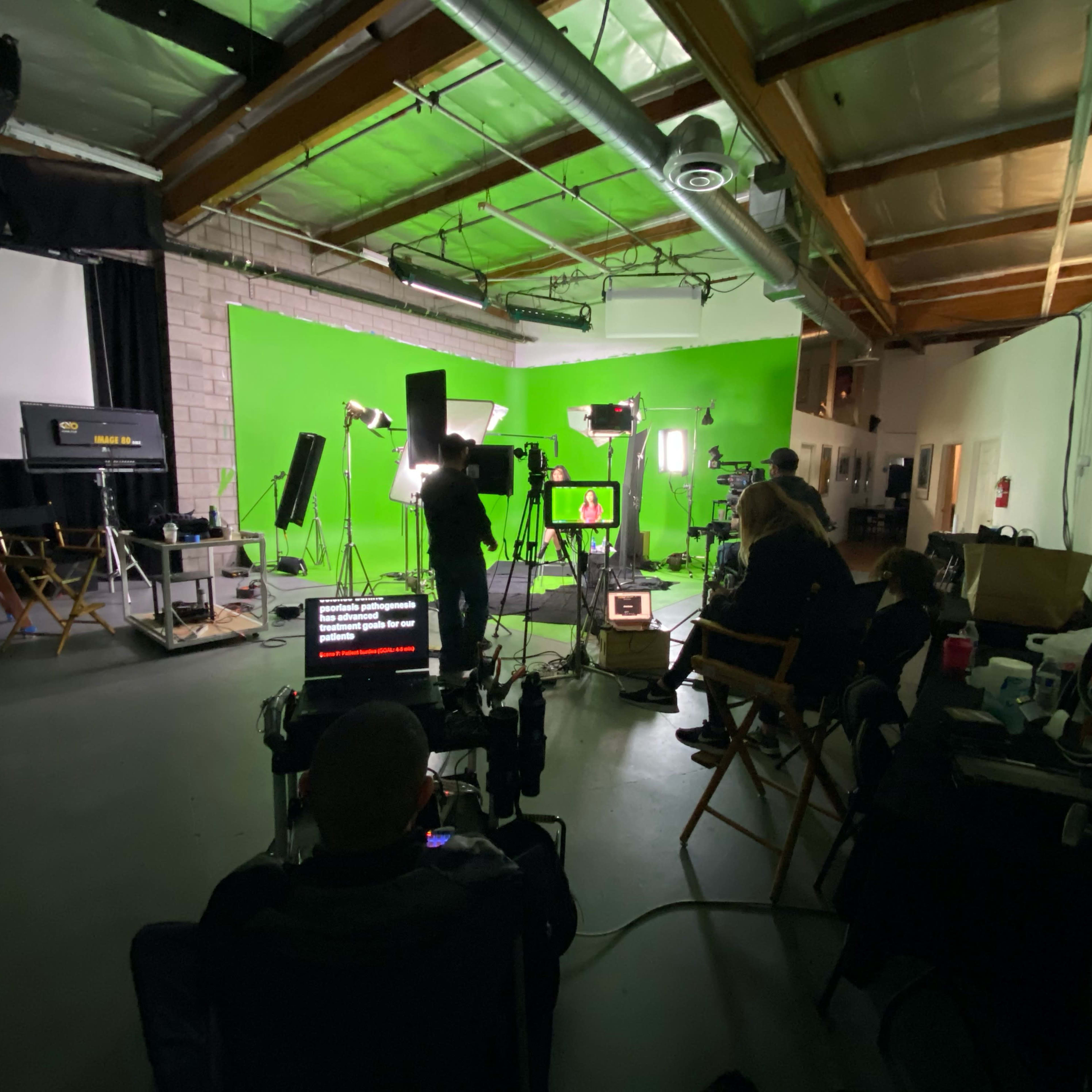 A film crew is set up in a studio with a green screen background, multiple lights, and equipment arranged for a shoot.