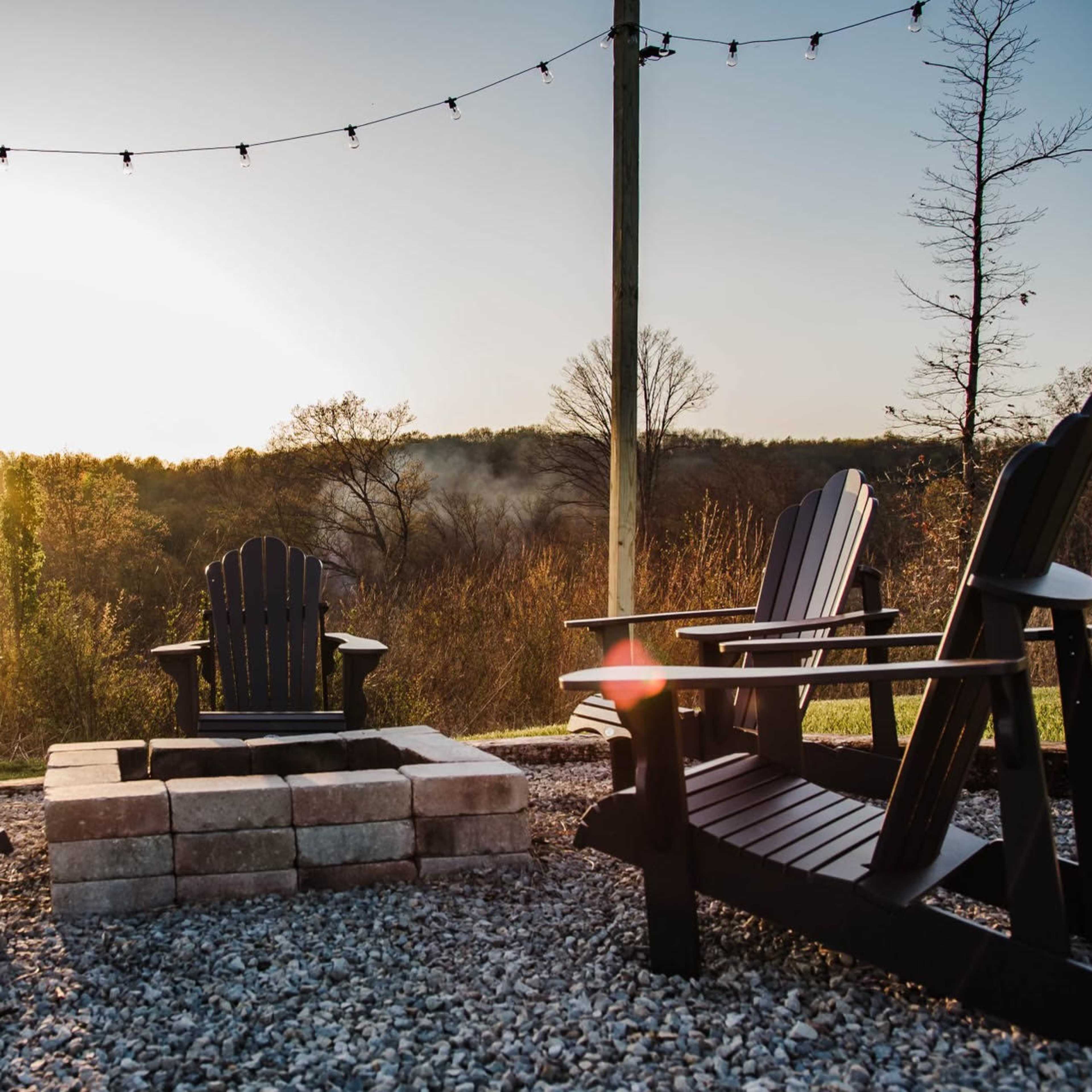 A circular arrangement of black wooden chairs surrounds a stone fire pit, set against a backdrop of trees and a distant sunsetting sky.