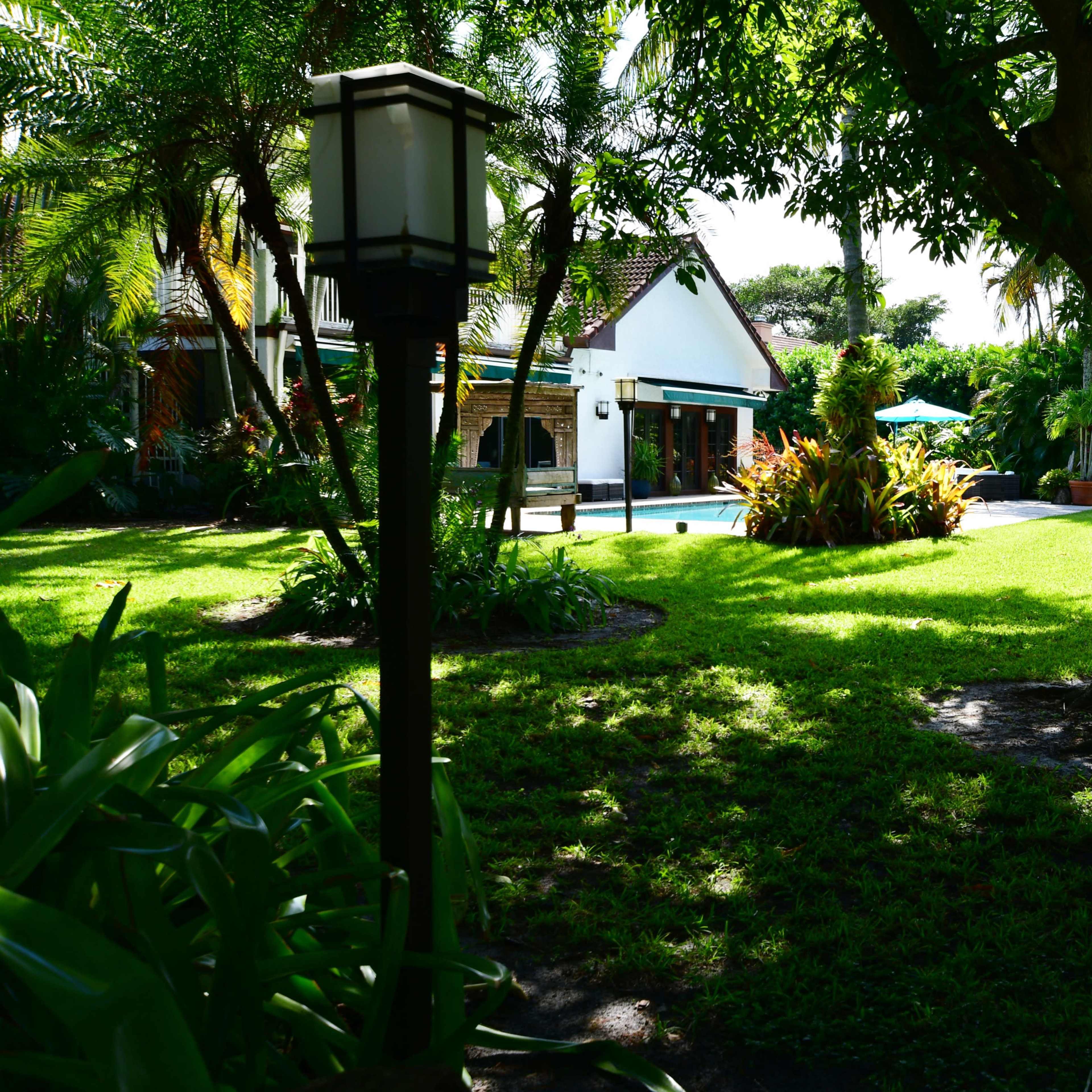 A spacious backyard features a well-maintained lawn, tropical plants, and a house with a pool visible in the background.