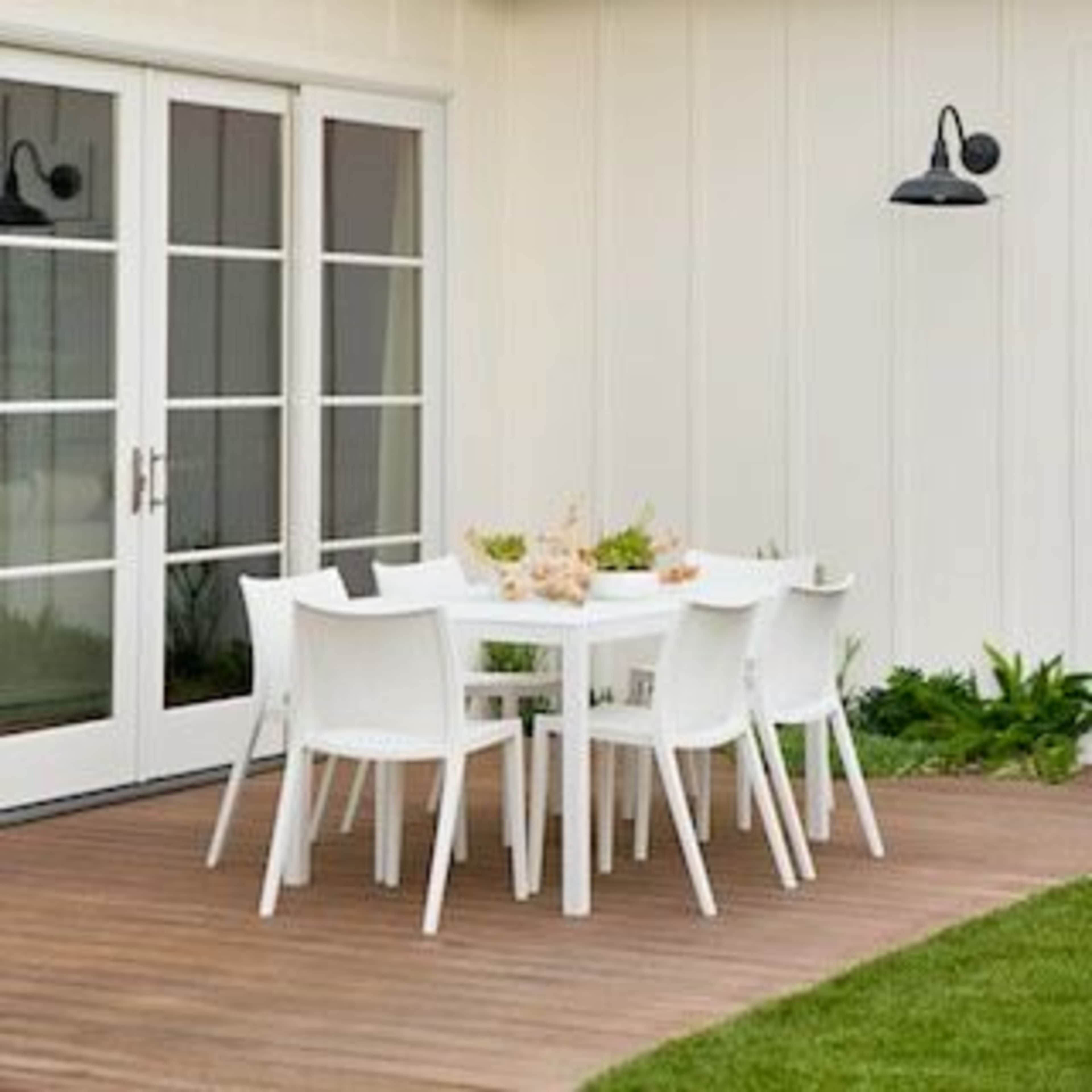 A modern outdoor dining area features a white table surrounded by white chairs on a wooden deck near a light-colored wall.