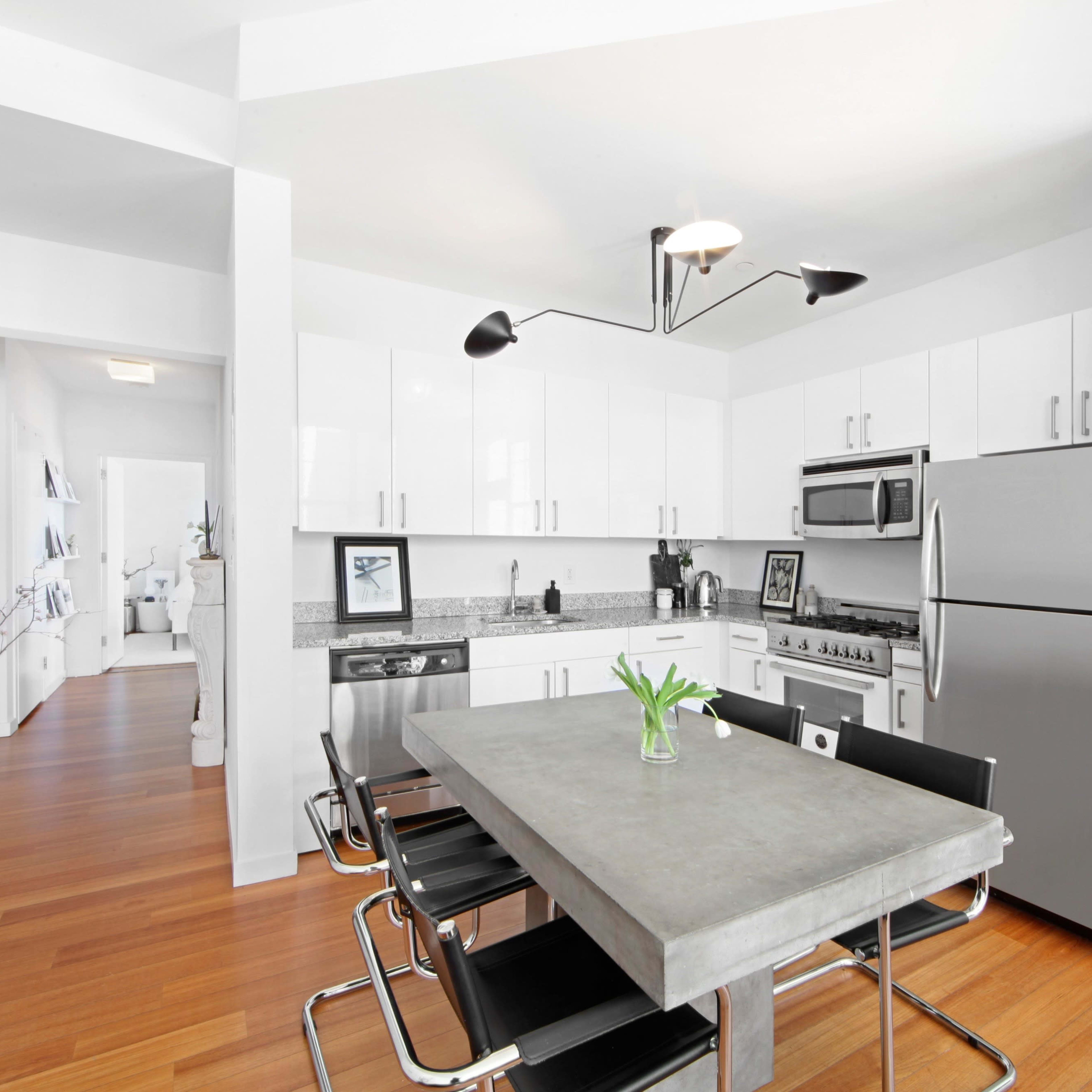 A modern kitchen with sleek white cabinets, stainless steel appliances, and a concrete dining table surrounded by black chairs.