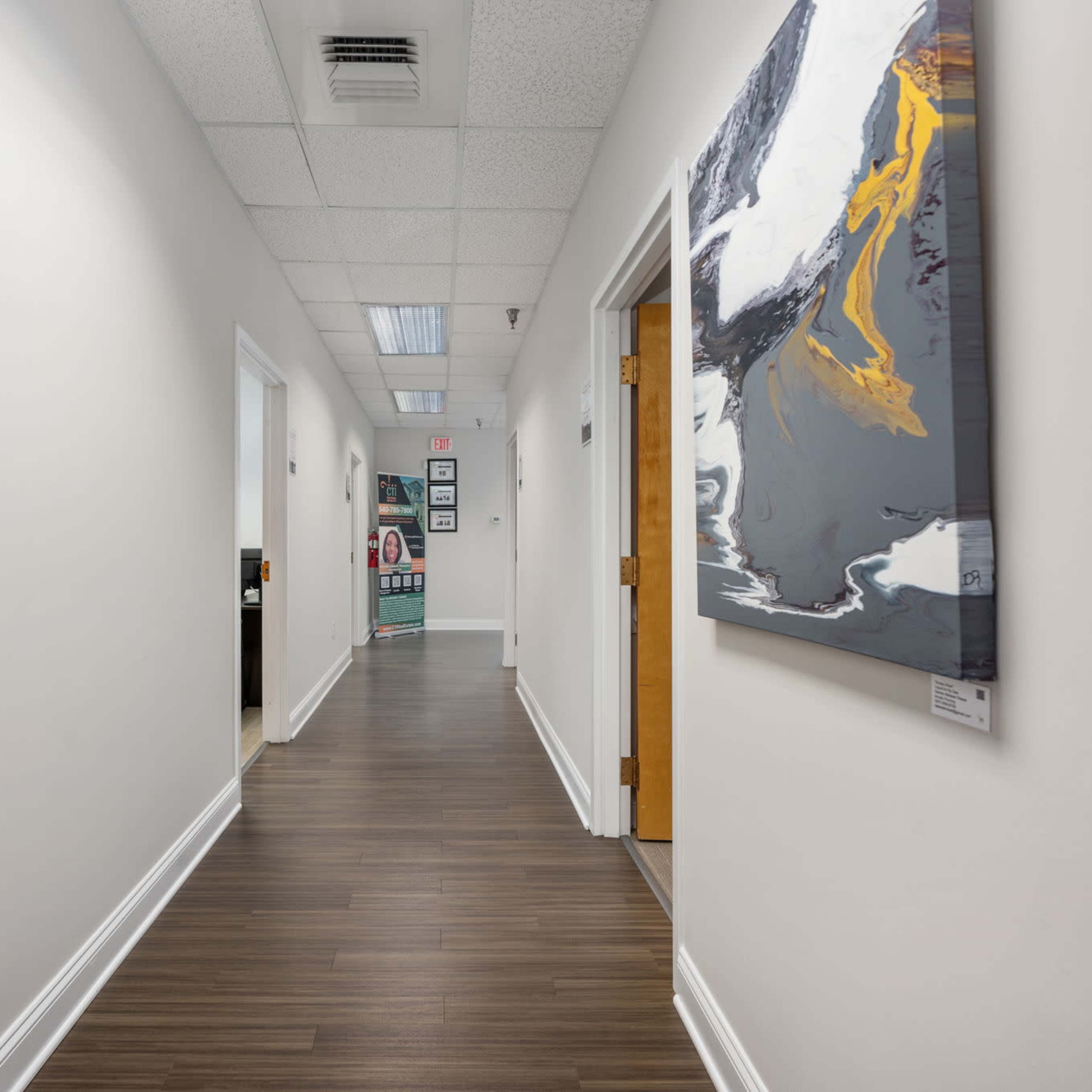 A hallway with wooden flooring, white walls, and doorways on either side, leading to an office space at the end.