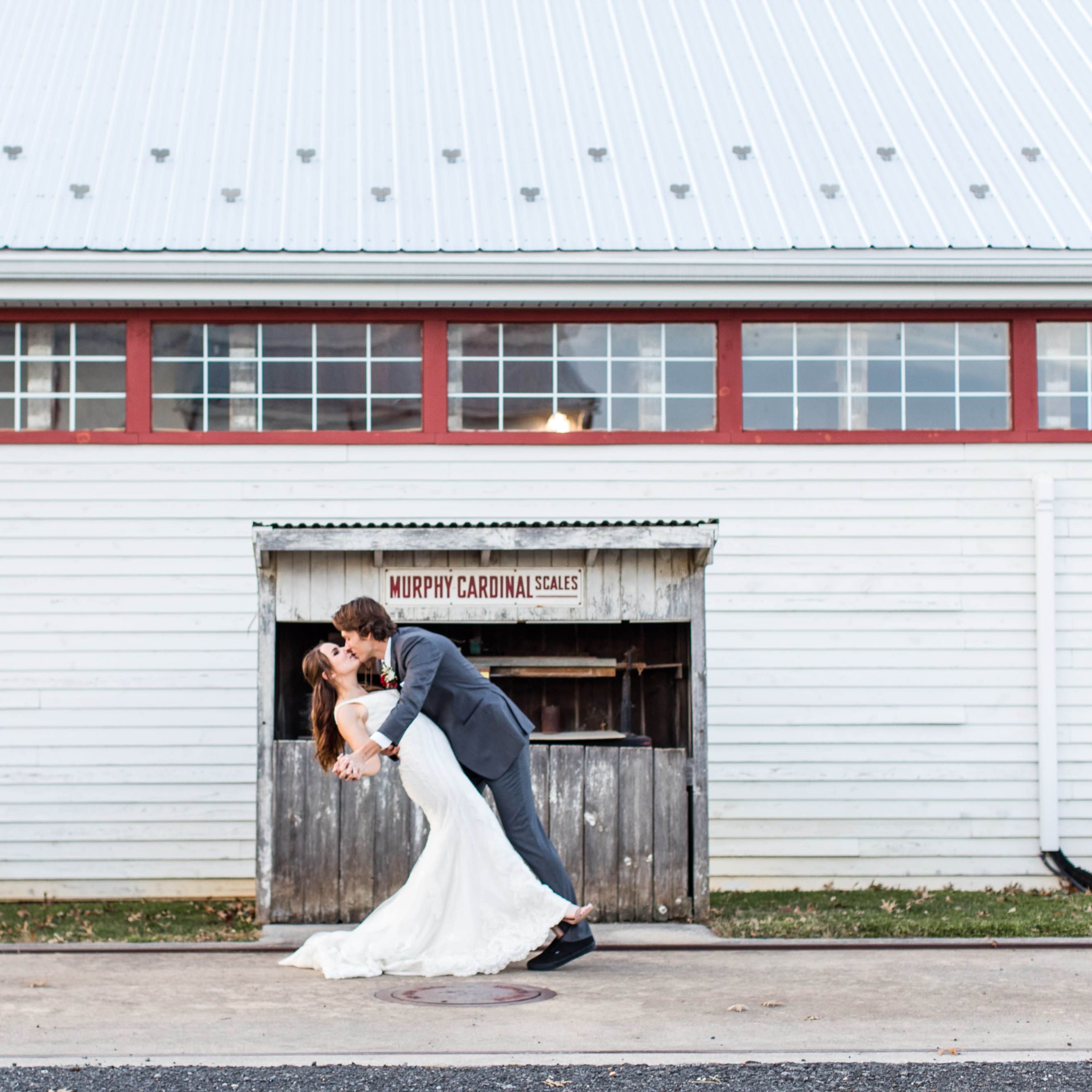 A couple in wedding attire is posed in a dip in front of a rustic barn with a sign reading "Murphy Cartina Scales."
