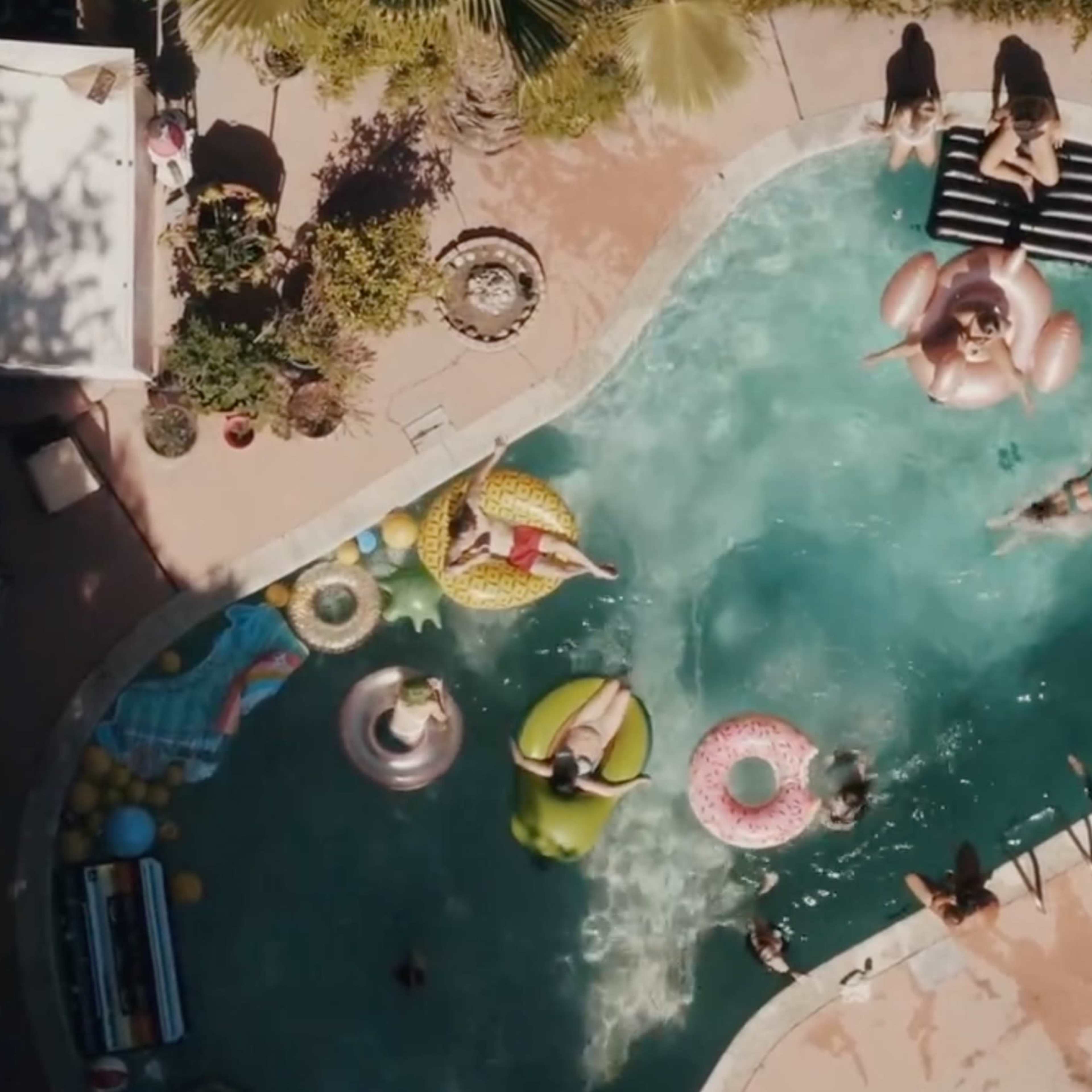 Aerial view of a swimming pool surrounded by lounge chairs, inflatables, and people enjoying leisure time.