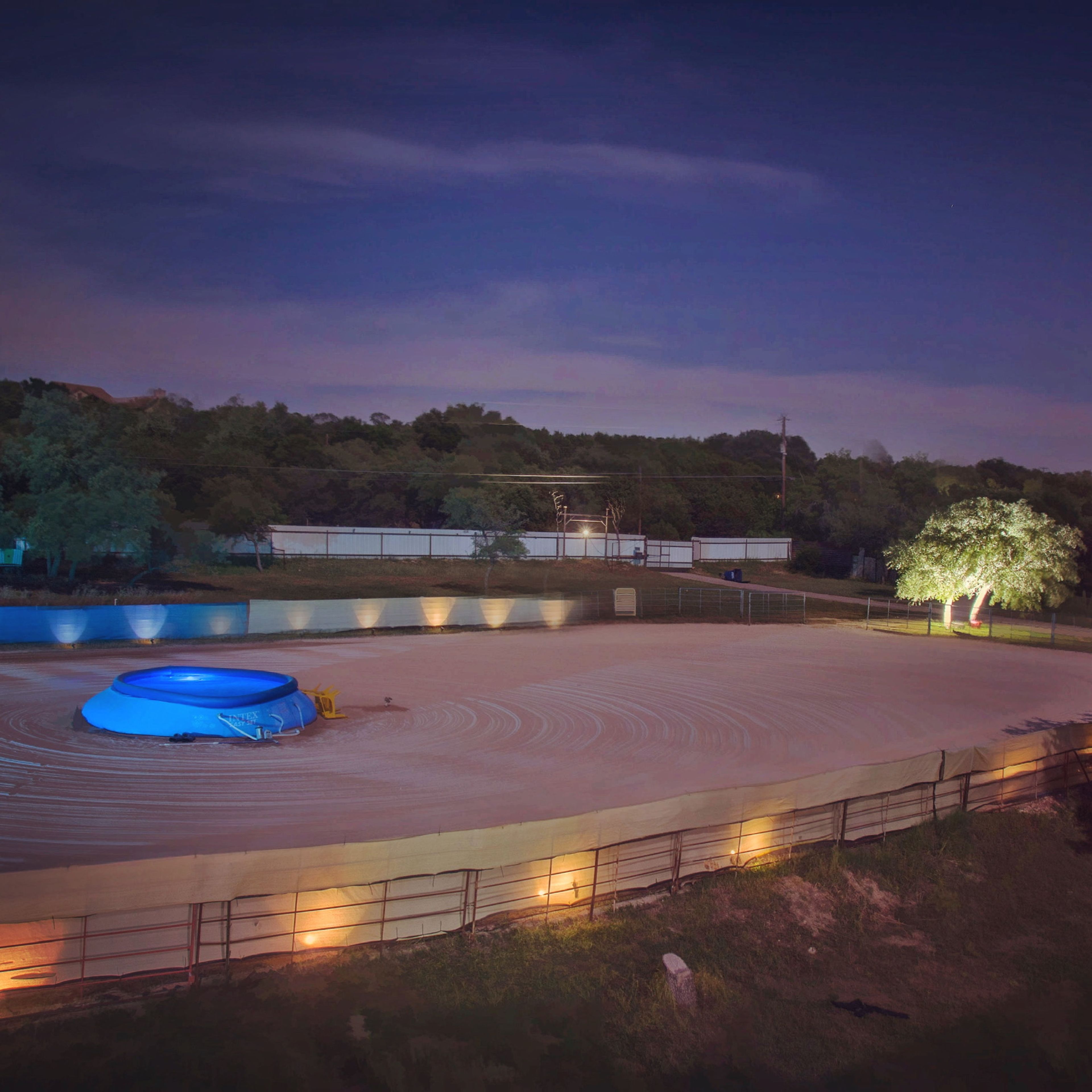 A blue circular above-ground pool sits in an empty, illuminated dirt area surrounded by trees at night.