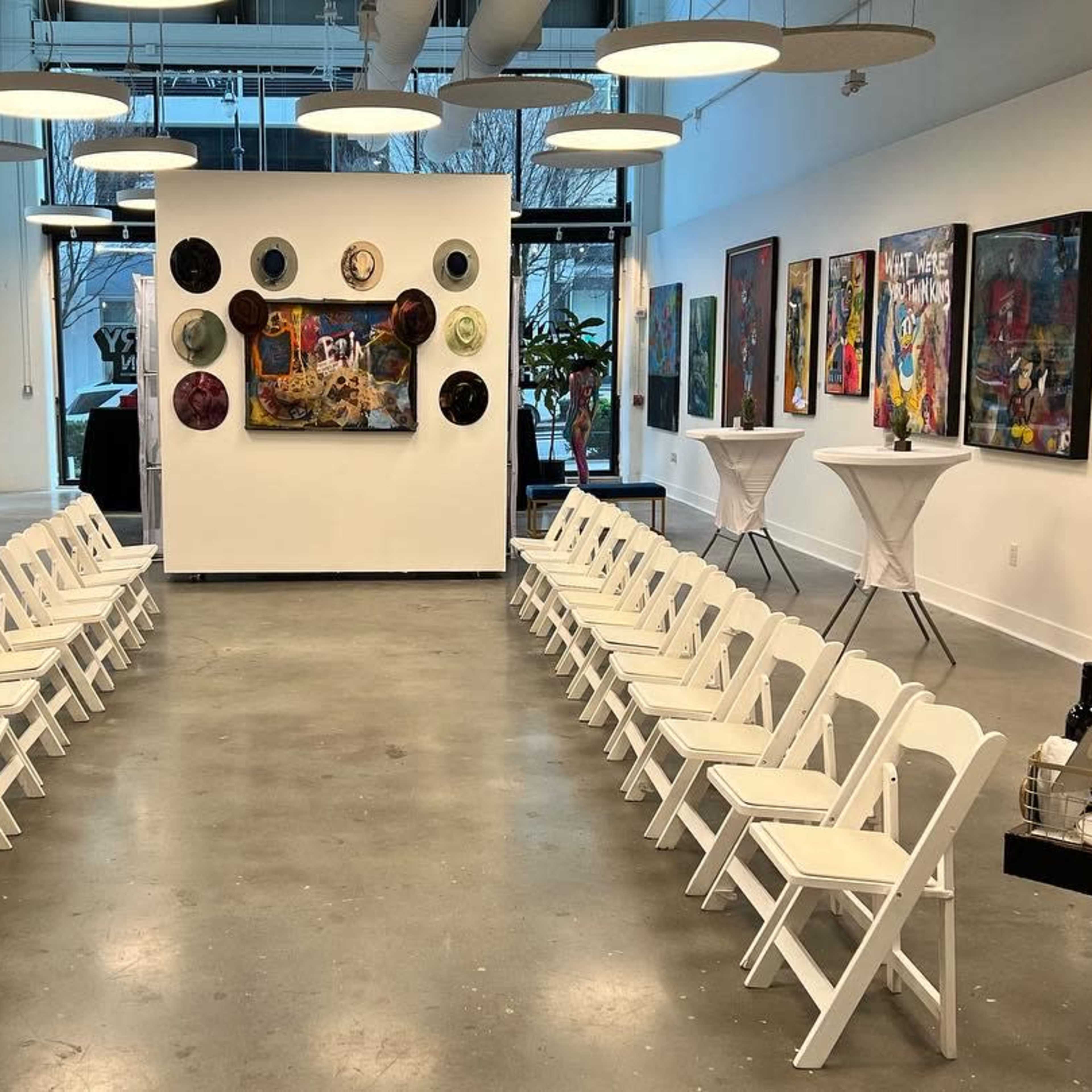An art gallery with rows of white folding chairs facing a central wall adorned with various artworks, surrounded by additional pieces on the side walls.