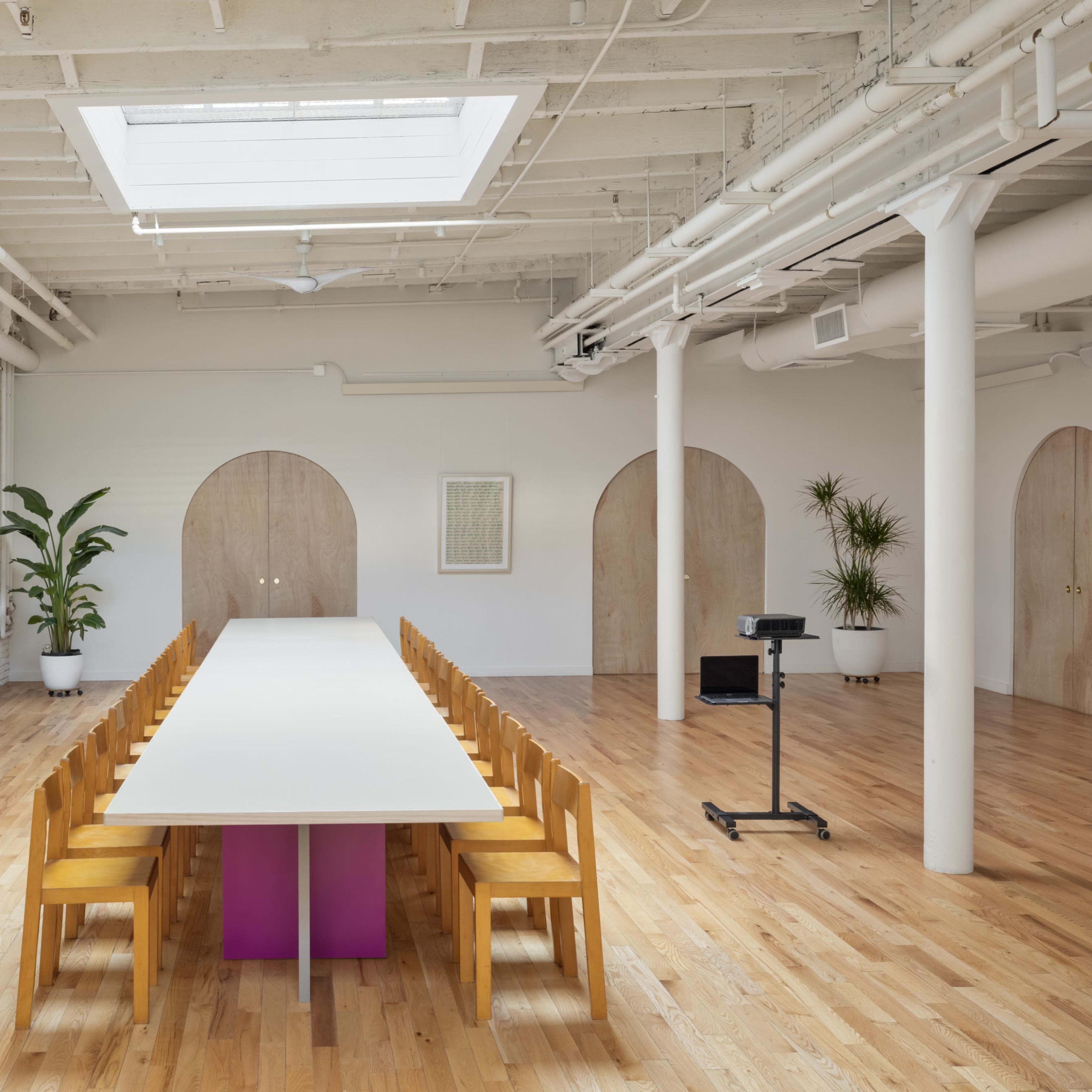 A bright meeting room features a long rectangular table surrounded by wooden chairs, with a projector screen and plants against the walls.