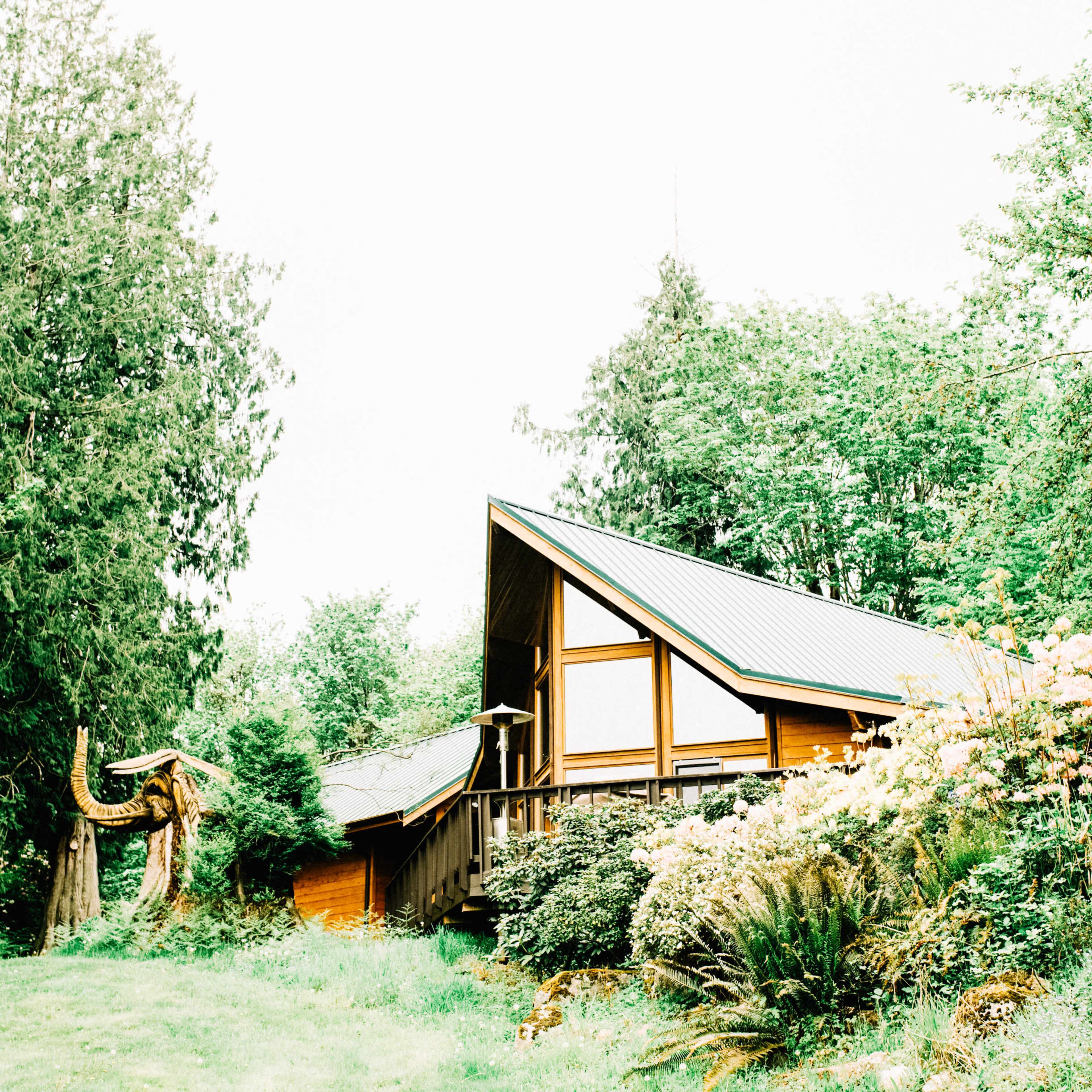 A wooden house with a triangular roof is surrounded by lush greenery and colorful flowers.