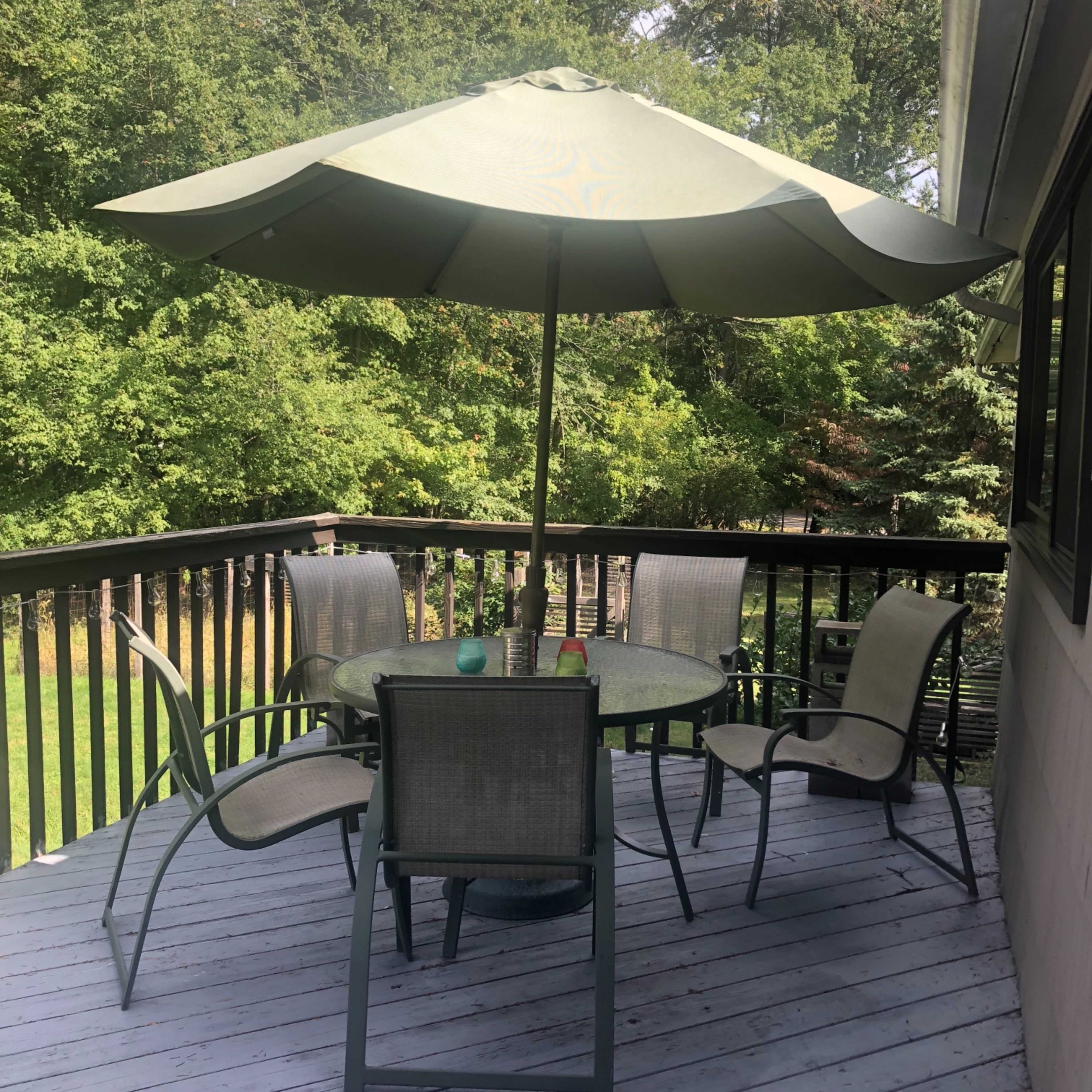 A round table with four chairs is situated under a green umbrella on a wooden deck surrounded by trees.