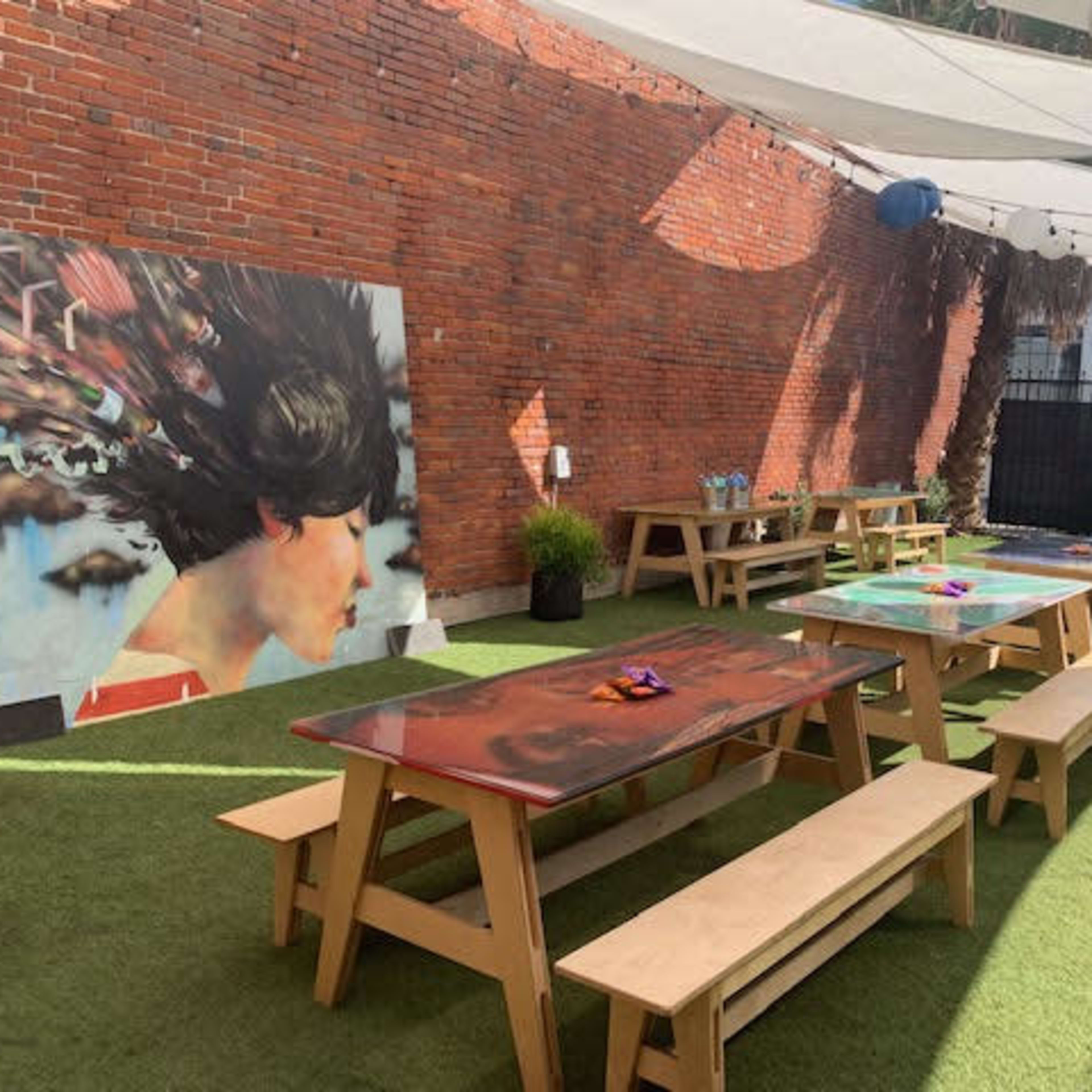 A courtyard with wooden picnic tables, a mural on a brick wall, and decorative hanging lanterns under a canopy.