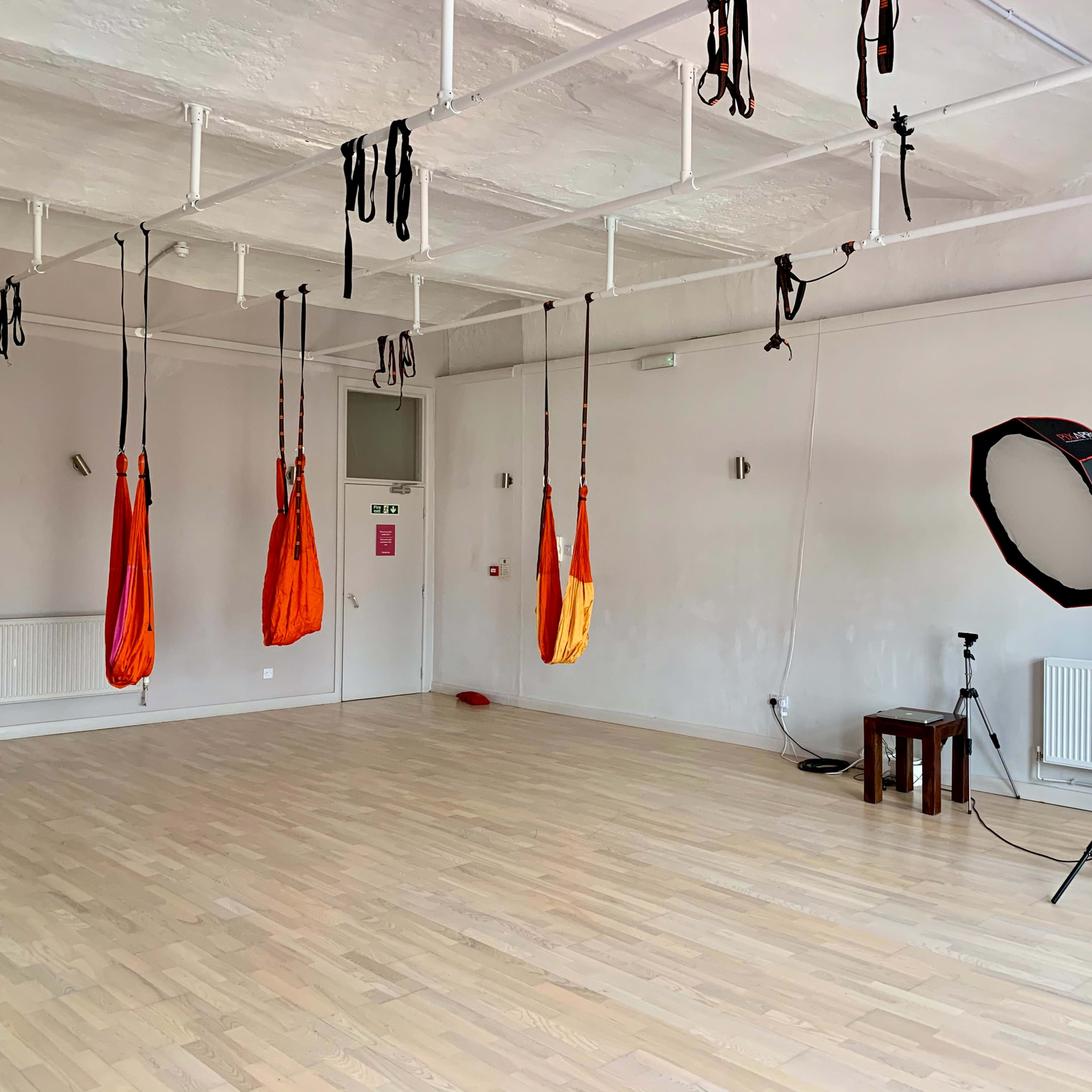 A spacious, empty studio features hanging aerial silks and a lighting setup in one corner.