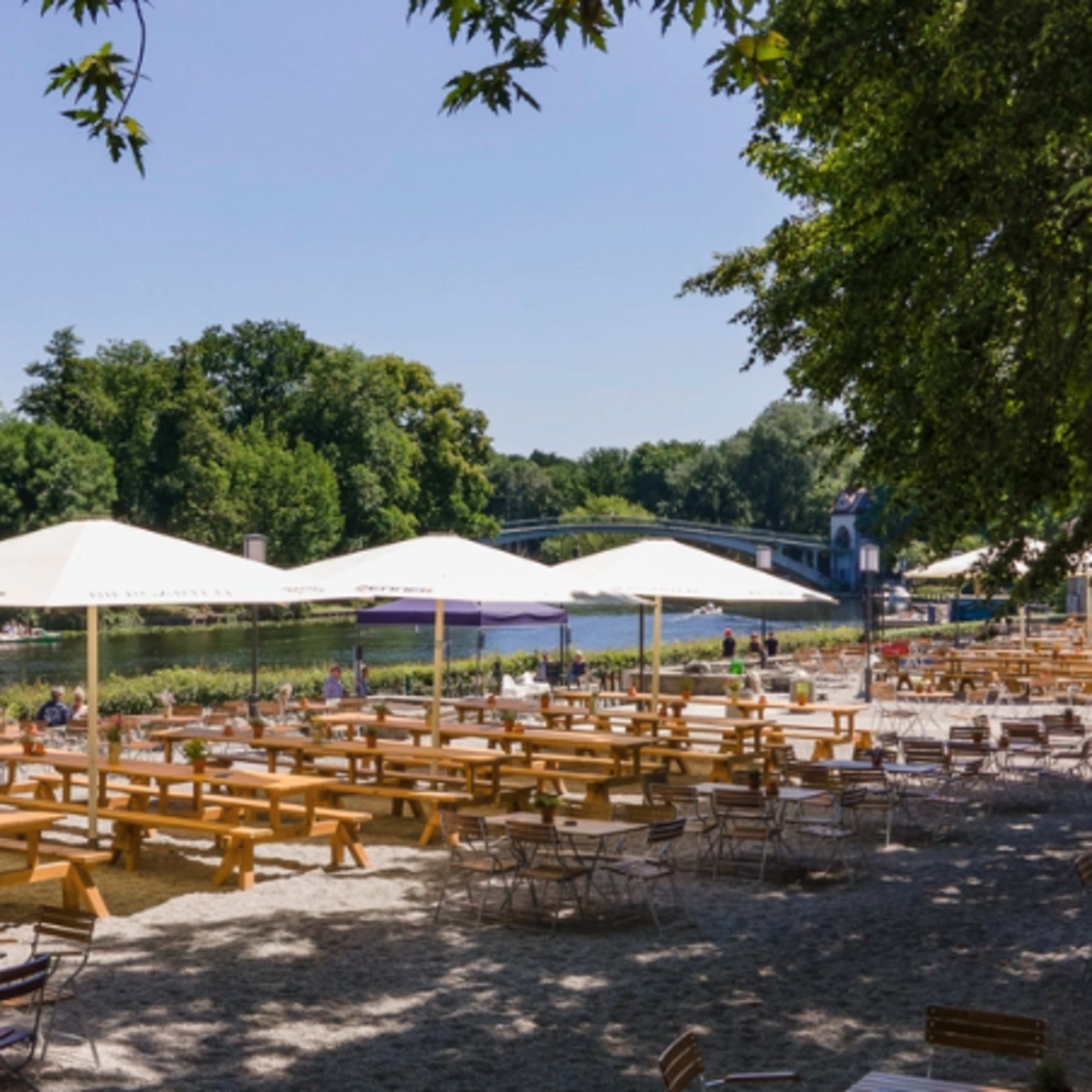 A riverside area features rows of wooden tables and chairs under large umbrellas, with trees lining the bank and boats visible on the water.