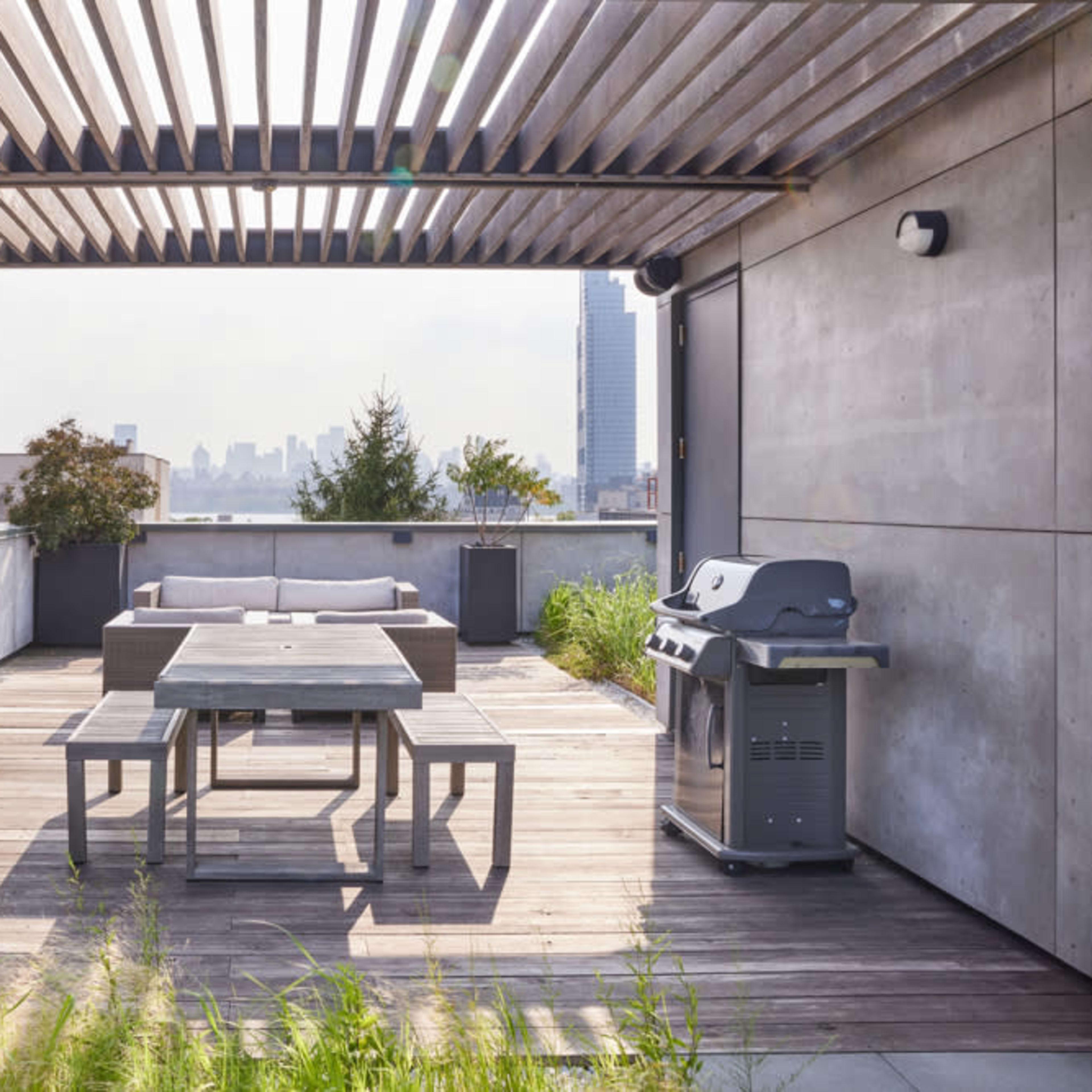 A modern rooftop terrace with wooden decking, potted plants, a dining table, and a grill, overlooking a city skyline.