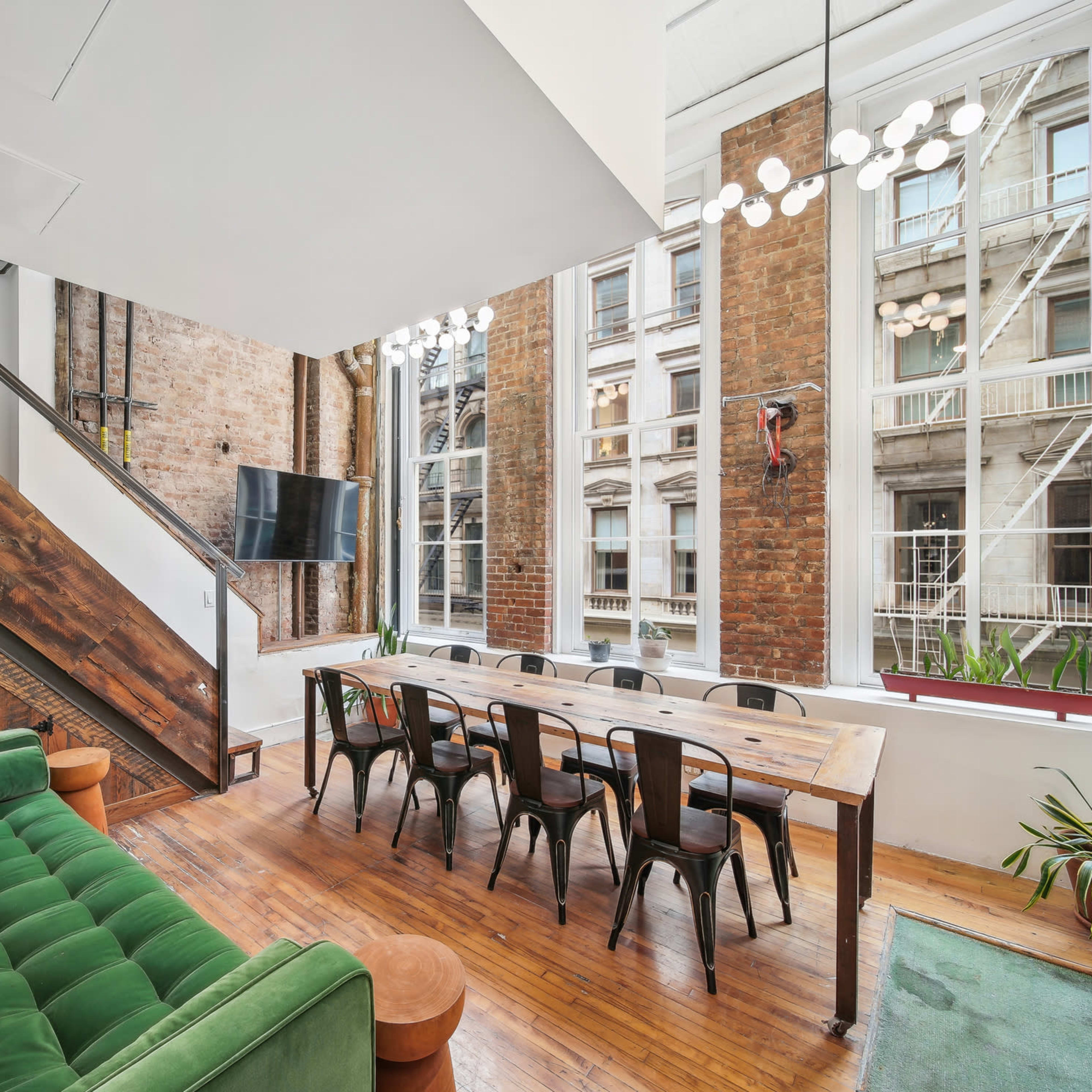 A modern interior features a green couch, a wooden dining table surrounded by black chairs, and large windows overlooking a brick building exterior.