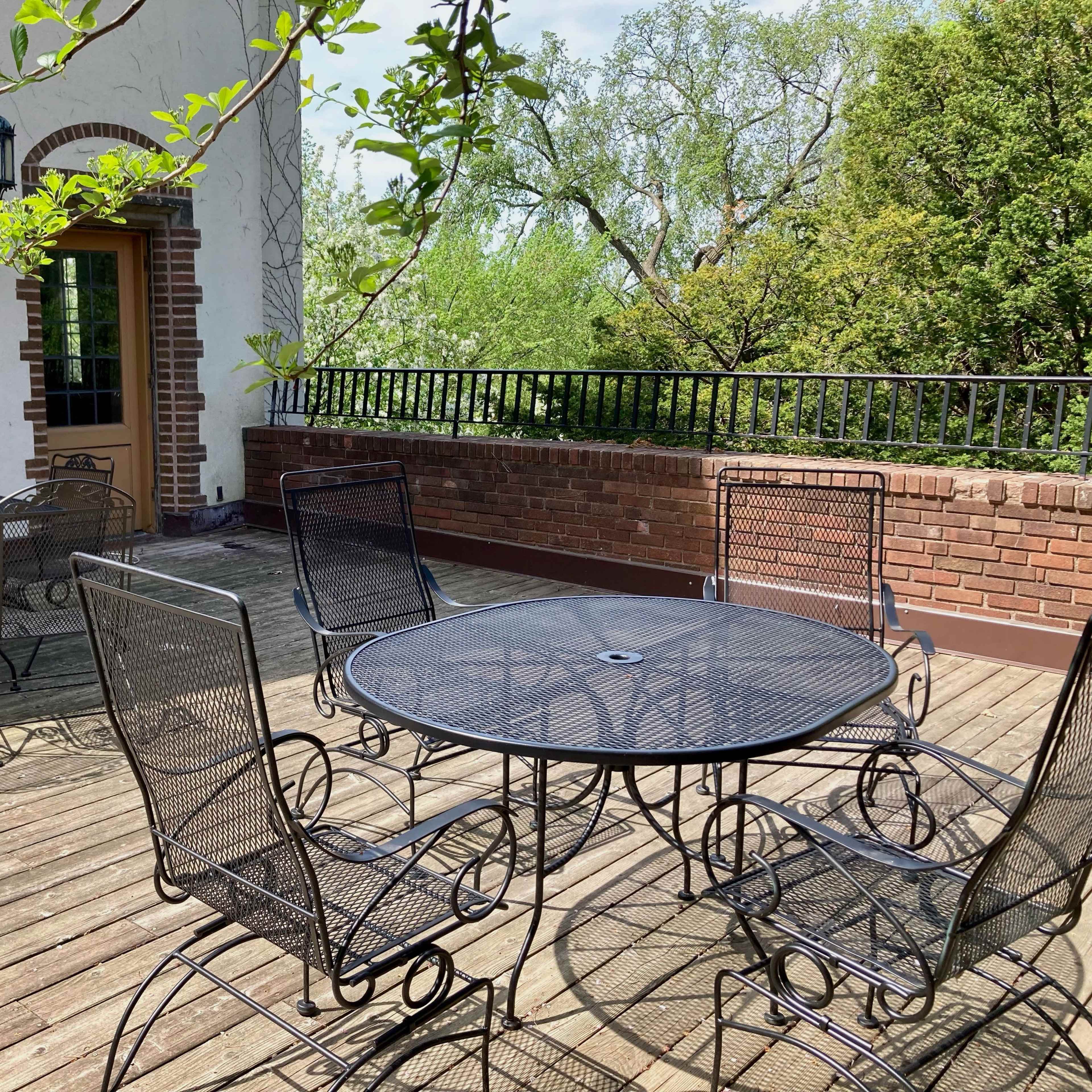 A spacious outdoor patio featuring several black wrought iron tables and chairs arranged on a wooden deck surrounded by greenery.