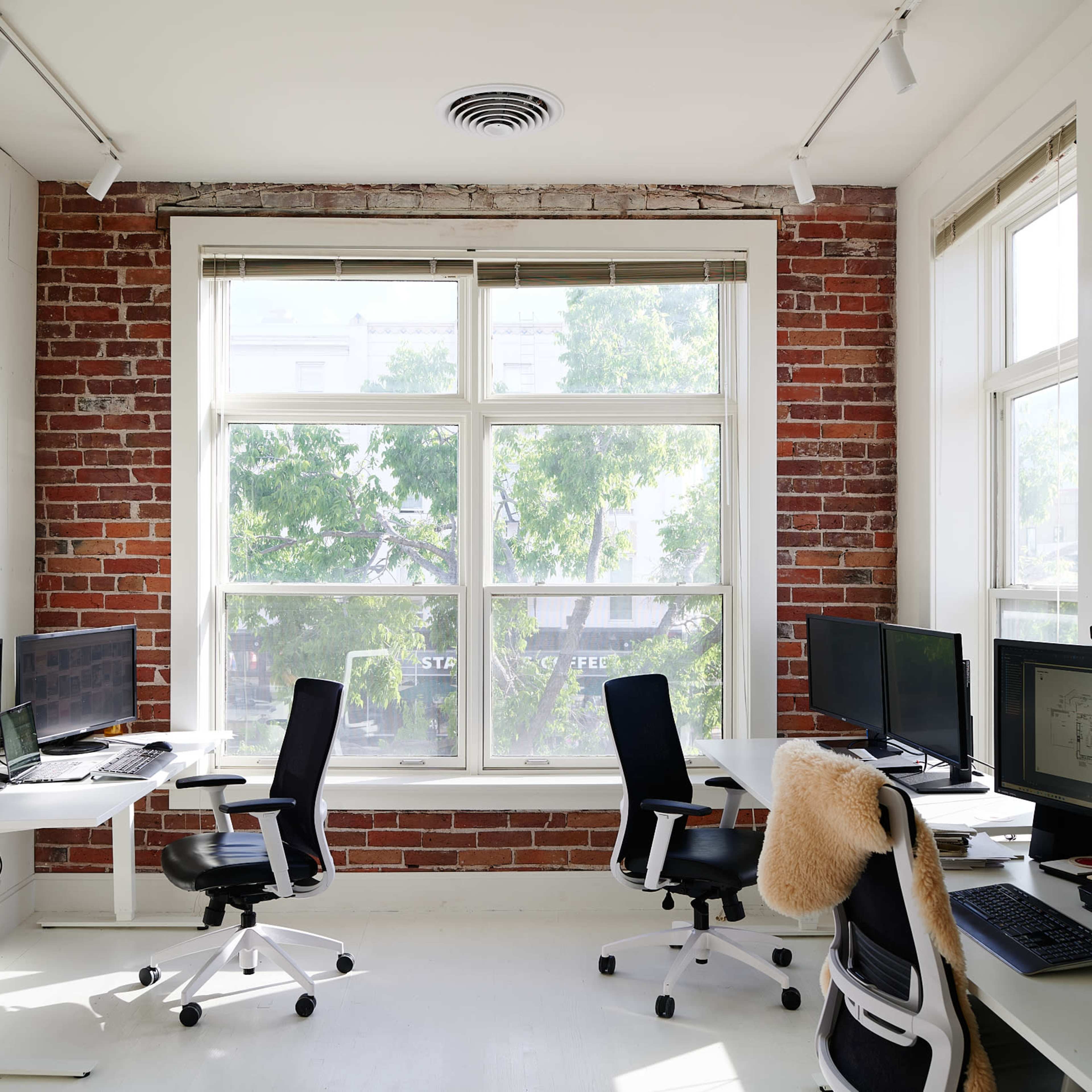 A bright office space with two workstations facing large windows, featuring exposed brick walls and modern furniture.